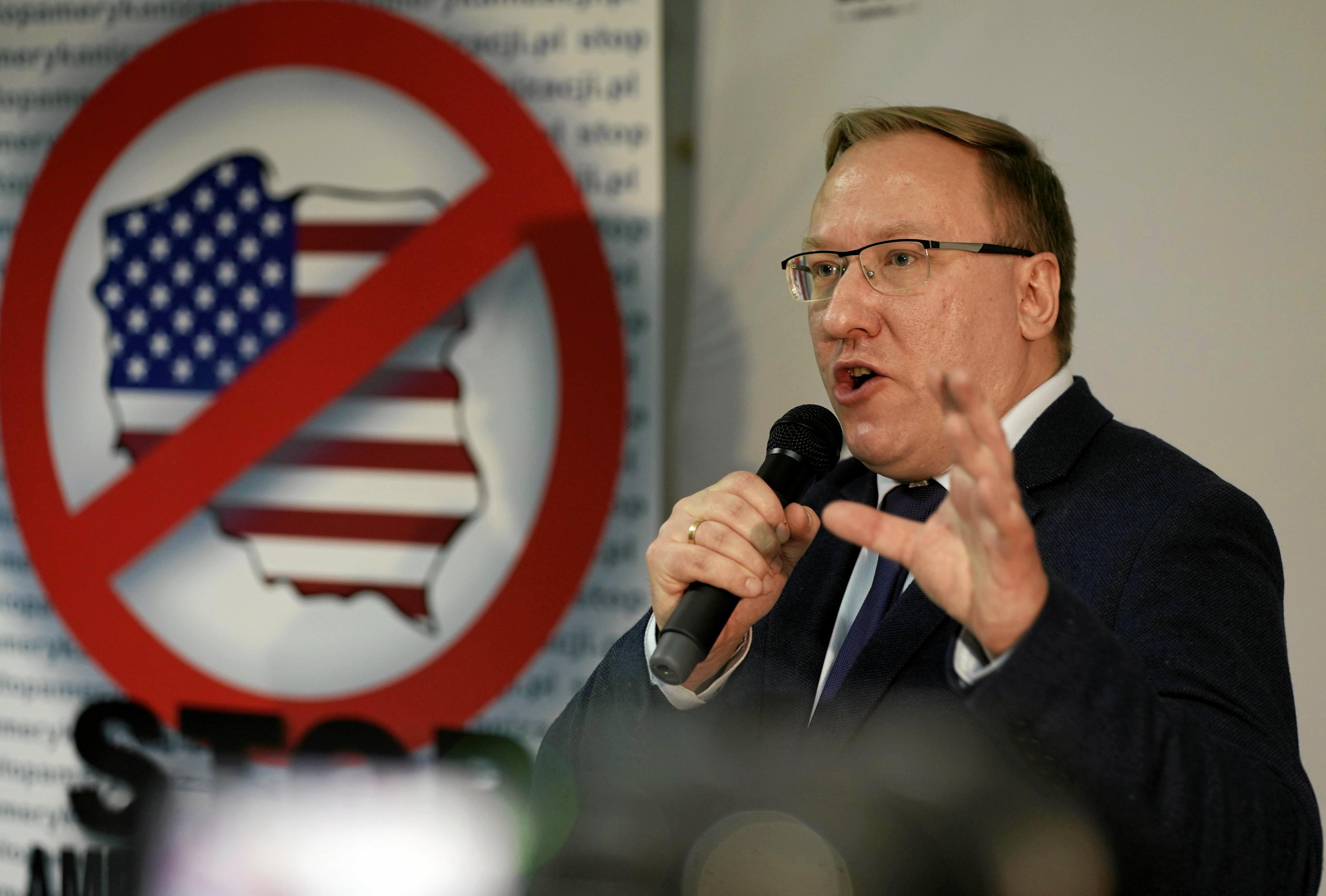 Mężczyzna w okularach mówi przez mikrofon na tle przekreślonej flagi Stanów Zjednoczonych