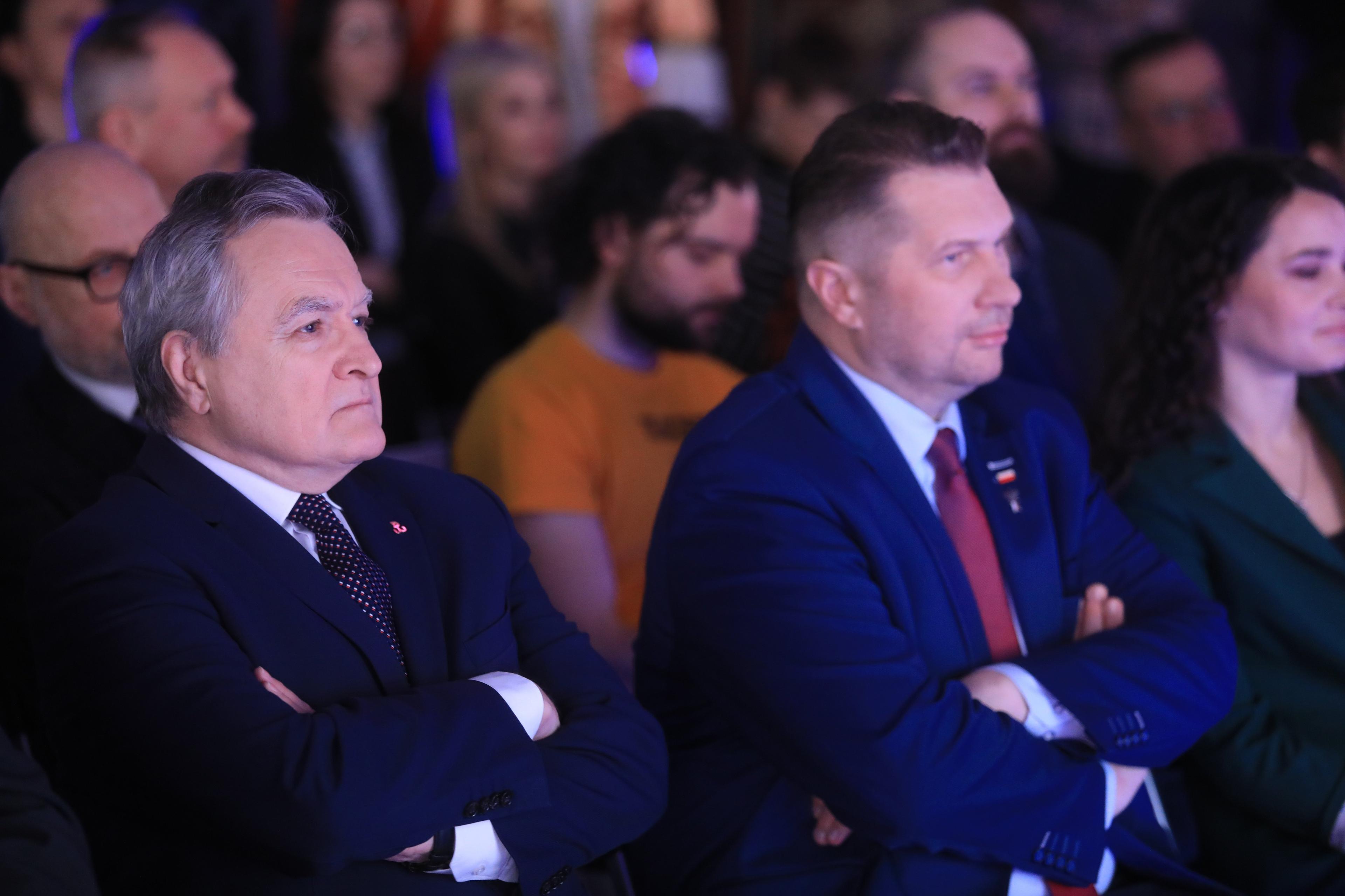 Dwaj mężczyźni - Piotr Gliński i Przemysław Czarnek - siedzą z założonymi rękami na sali pełnej ludzi