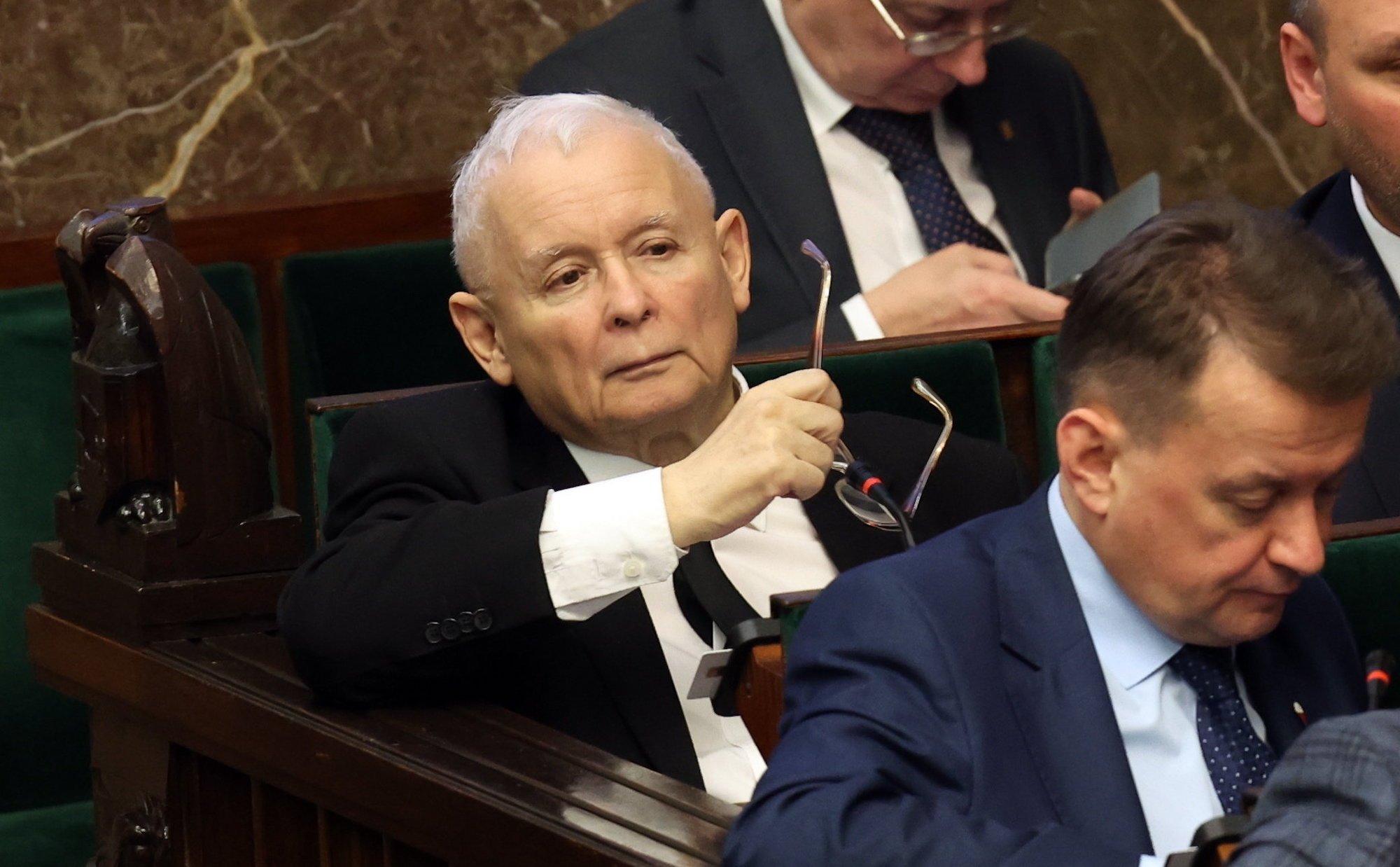 Jarosław Kaczyński w sejmowej ławie. Spogląda przed siebie z poważną miną, lekko unosząc brwi. W ręce trzyma okulary.