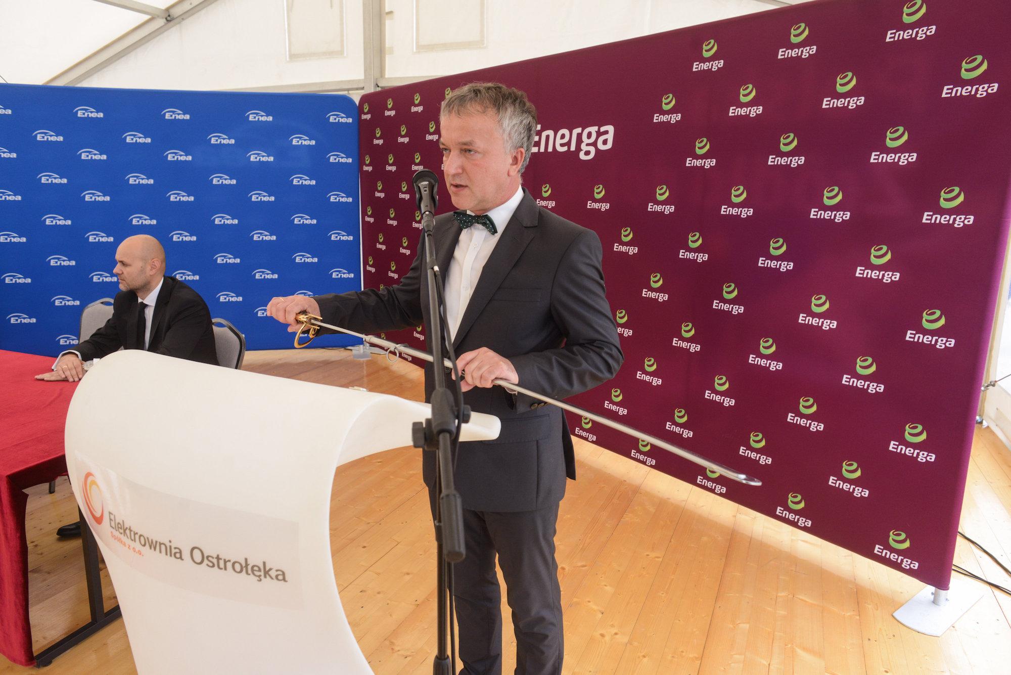 Ówczesny prezydent Ostrołęki Janusz Kotowski w smokingu i czarnej muszce przemawia na konferencji prasowej. W ręku trzyma szablę, stoi na tle ścianek z logo firm Energa i Enea.
