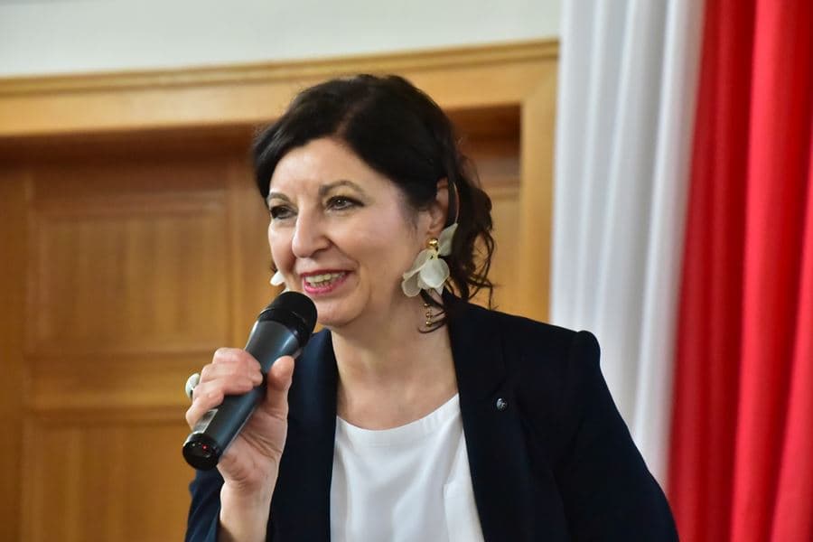 Kobieta z ciemnymi włosami, w eleganckiej marynarce przemawia do mikrofonu