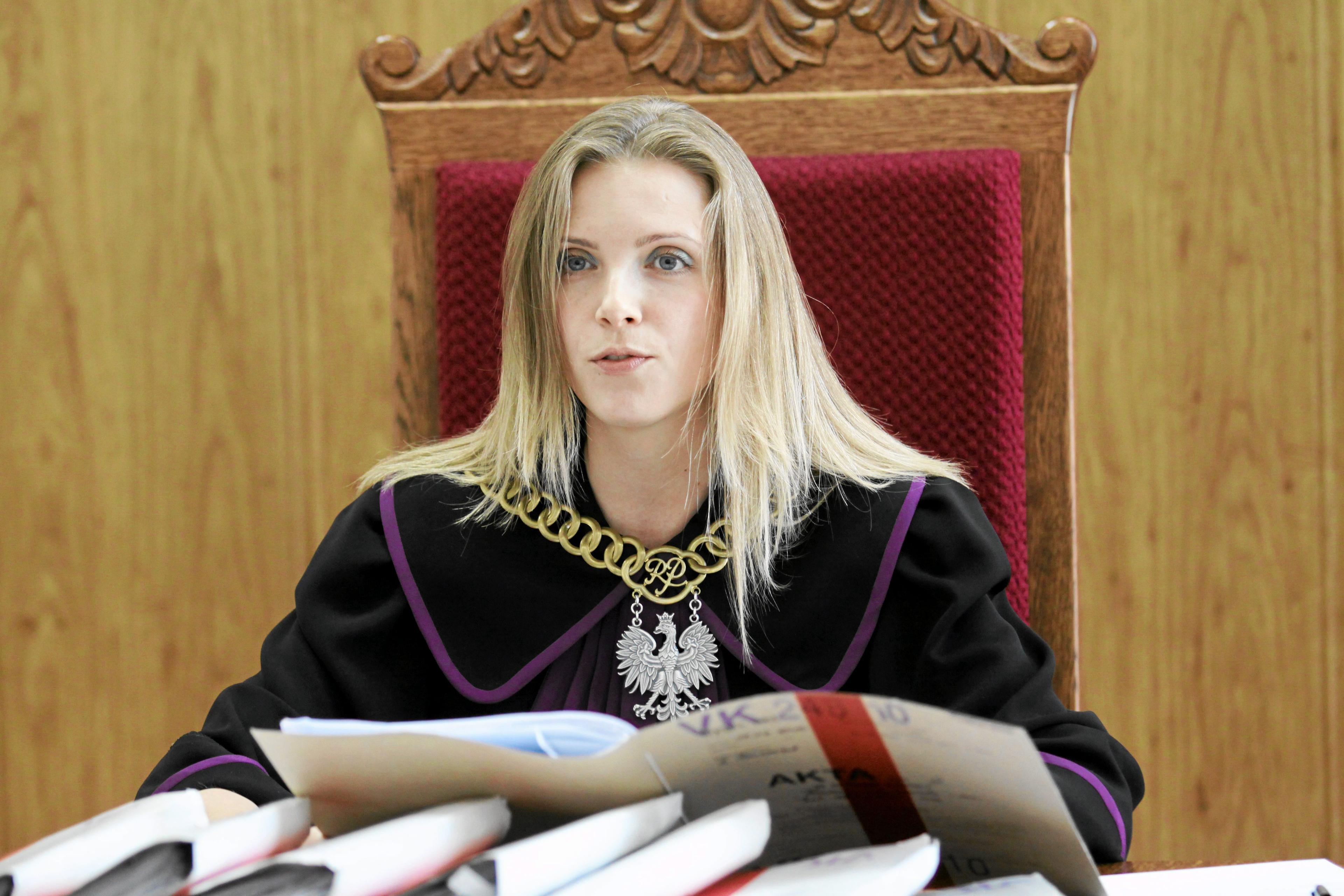 Portret sędzi Izabeli Szumniak. Kobieta w todze sędziowskiej i sędziowskim łańcuchy na szyi siedzi za stołem, na którym leżą rozłożone akta.