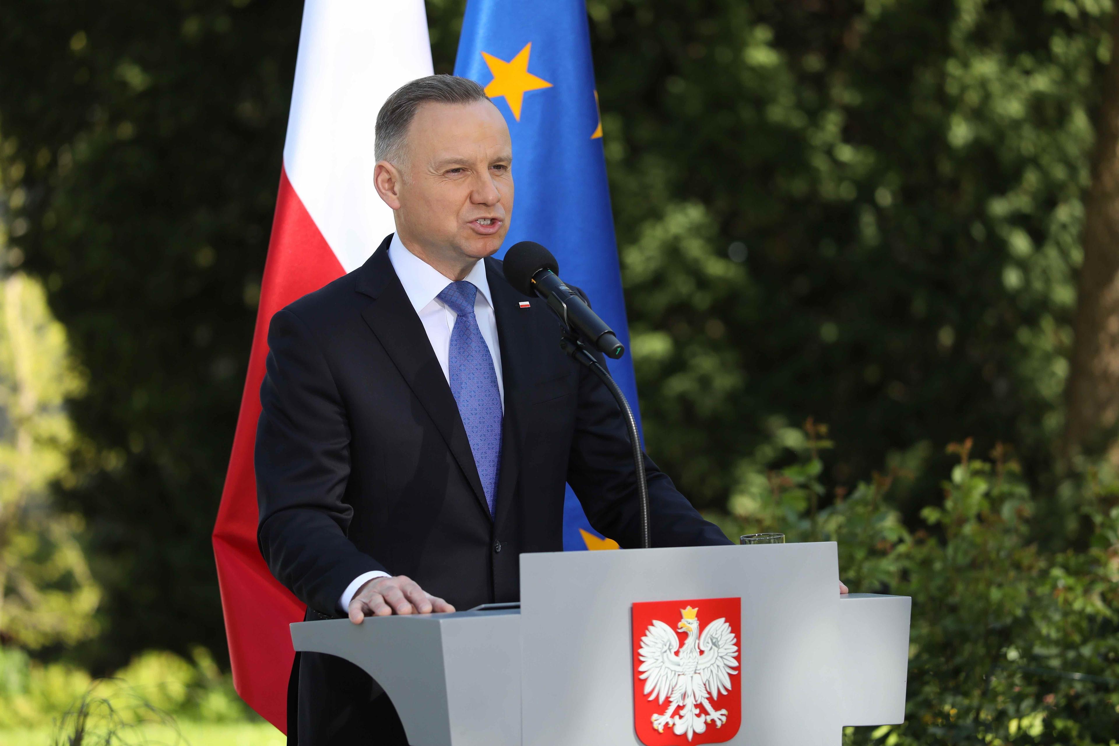 Prezydent Andrzej Duda przemawia w ogrodzie za mównicą z polskim godłem.
