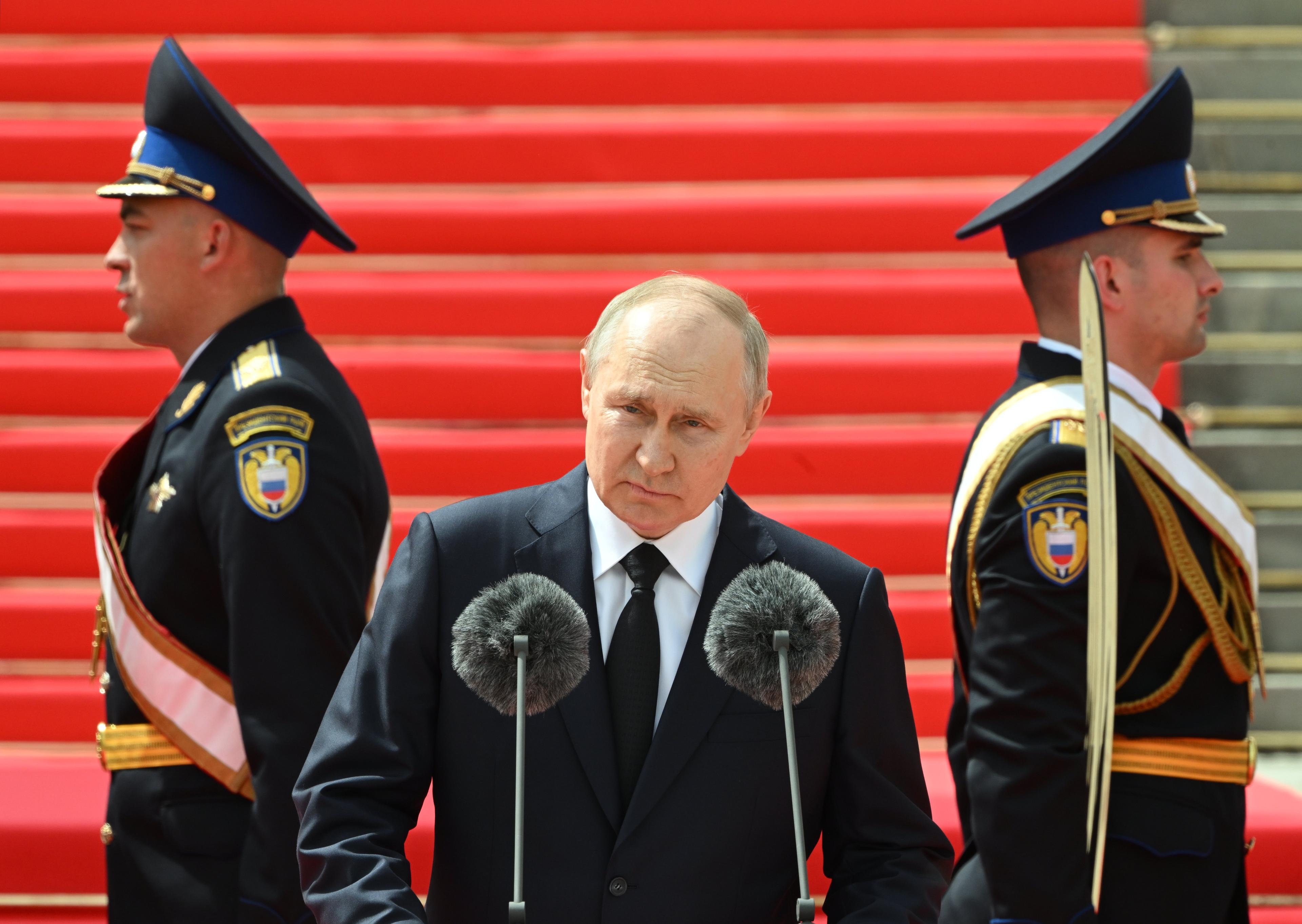 Putin przemawia, za nim dwaj żołnierze patrzący w dwie rózne stromy i zwróceni do siebie plecami
