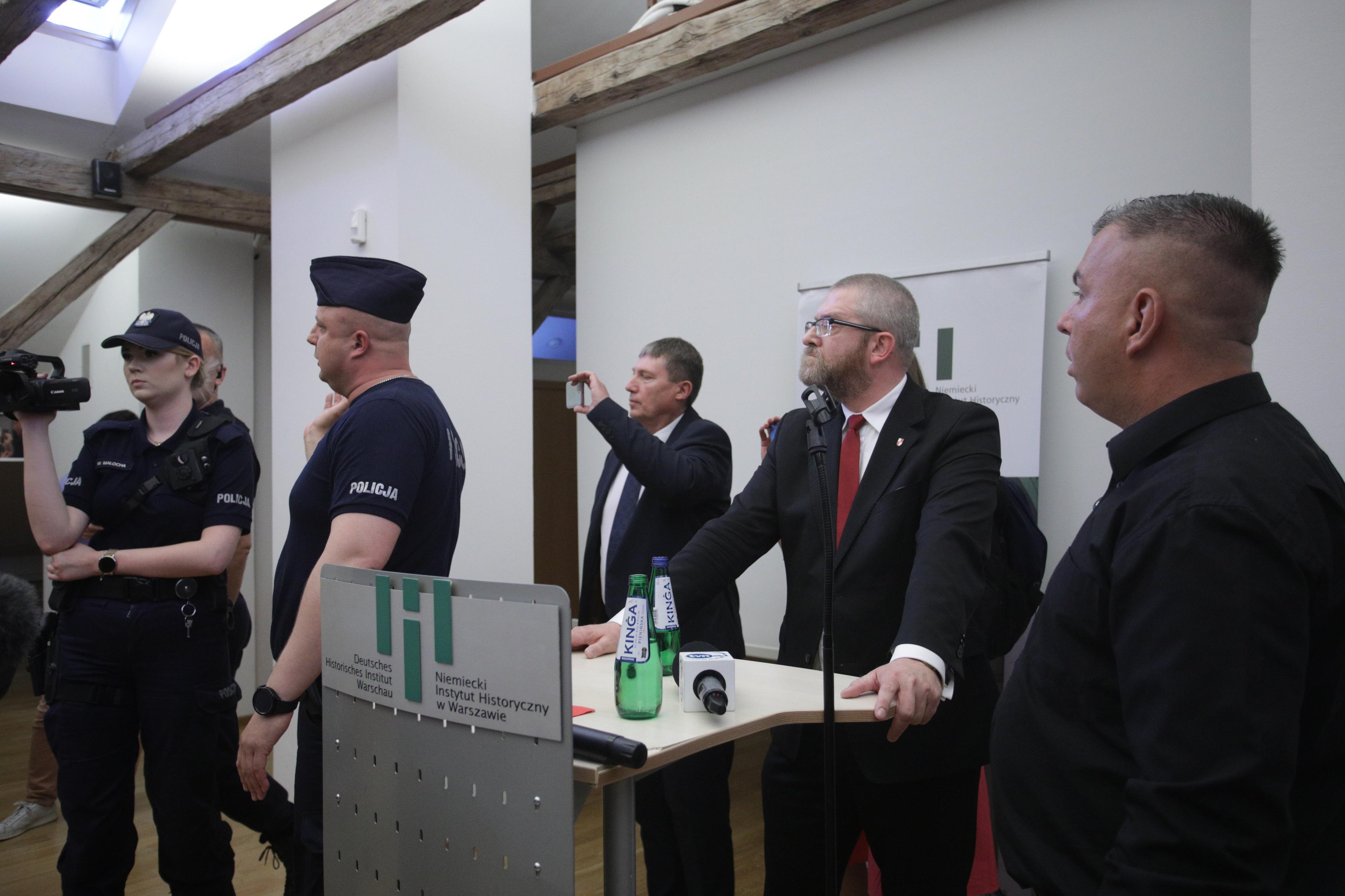 Poseł Grzegorz Braun (w okularach, z brodą) stoi przy mównicy, obok policjant i uczestnicy przerwanego wykładu prof. Grabowskiego