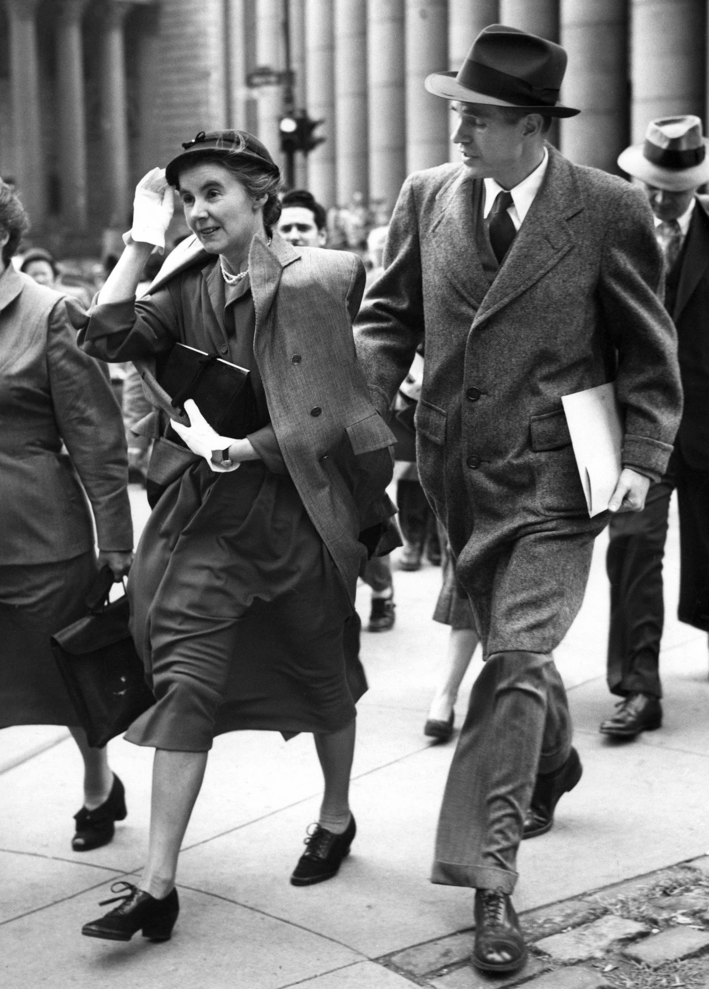 Czarno-białe zdjecie przedstawiające mężczyznę w płaszczi i kapeluszu i kobietę w garsonce, idących ulicą (Joseph MacCarthy).