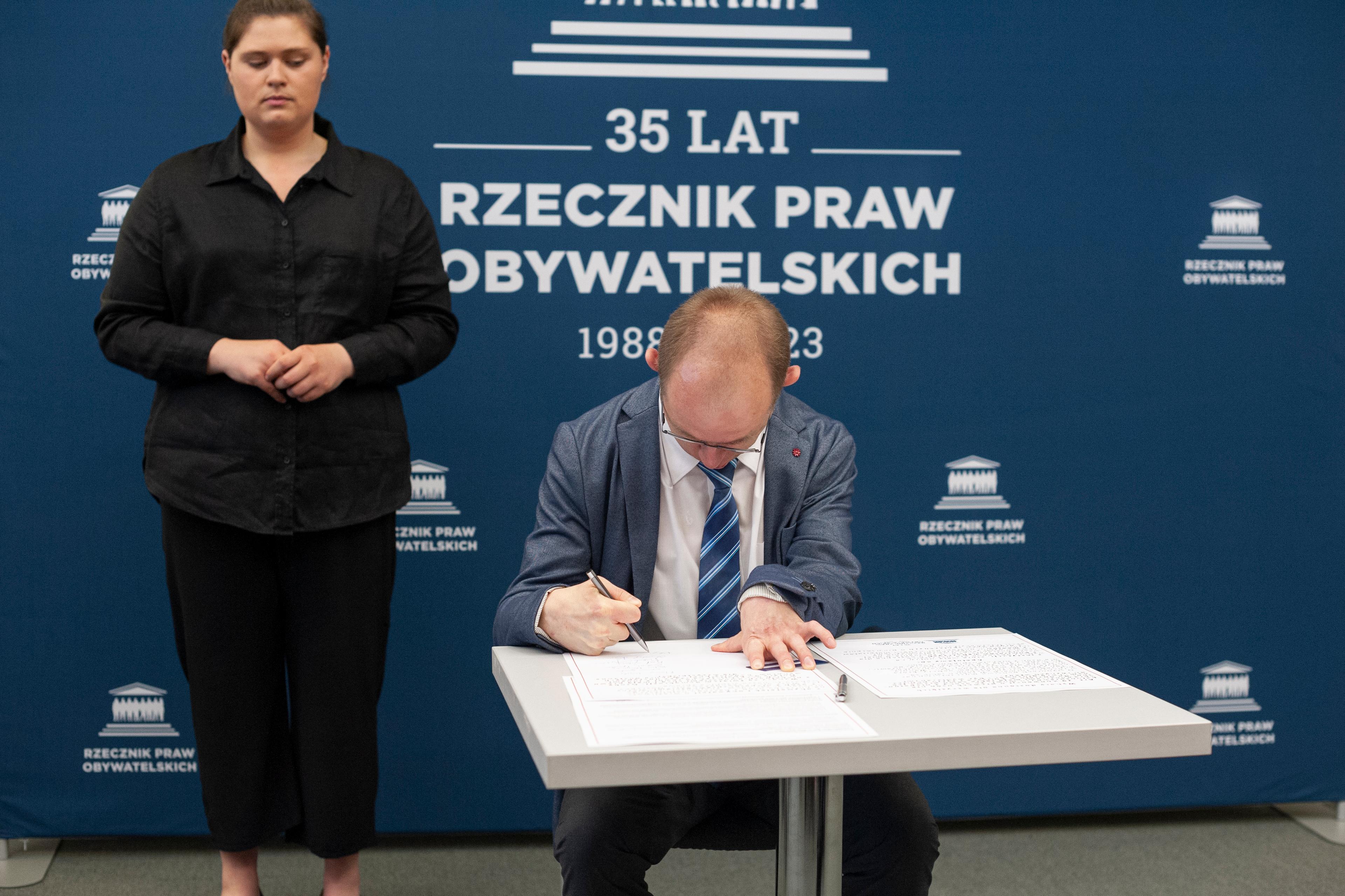 Mężczyzna w garniturze (Krzysztof Kurowski)odpisuje dokument. Obok - tłumaczka języka migowego