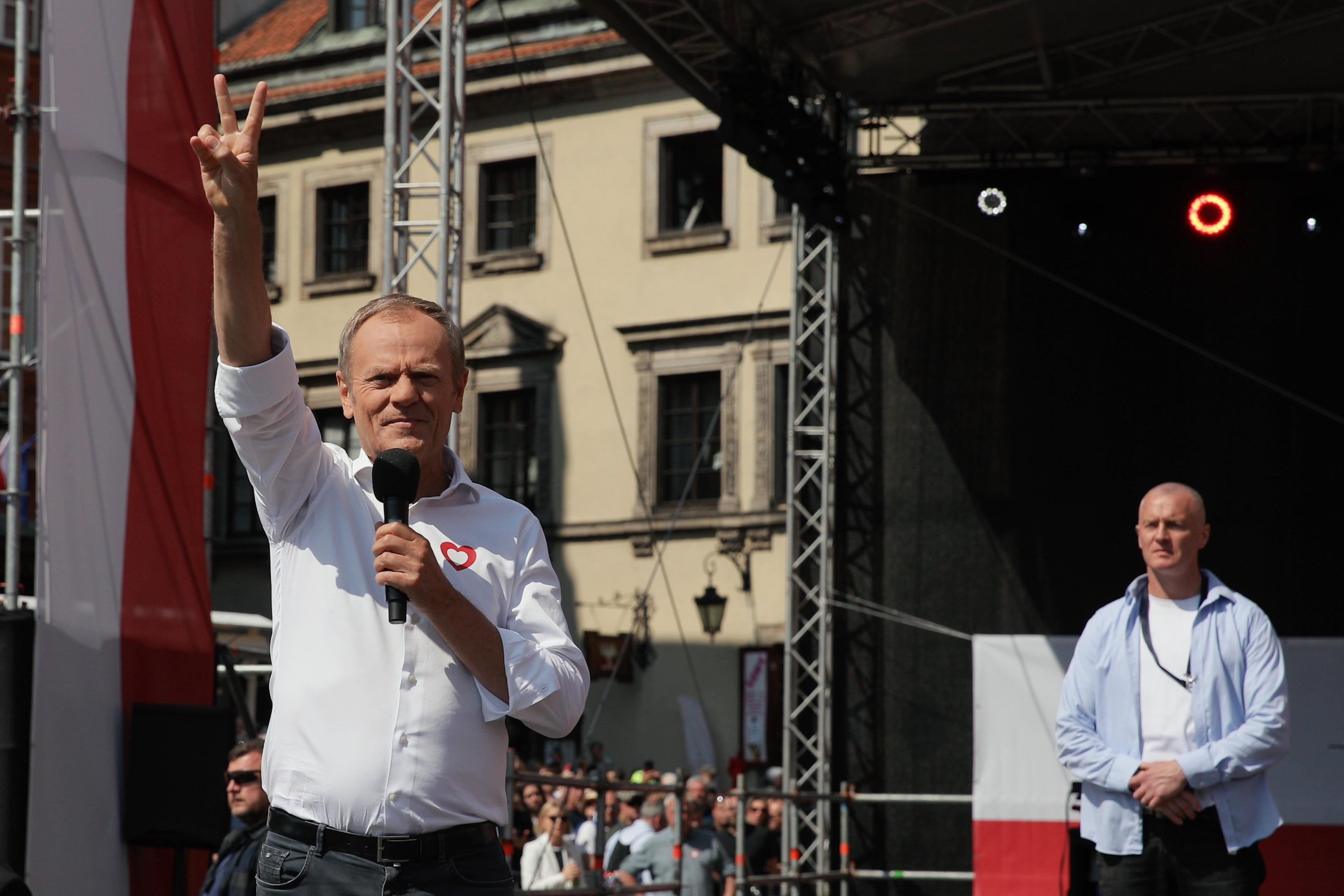 Mężczyzna stoi na scenie, w jednej dłoni trzyma mikrofon, drugą pokazuje znak zwycięstwa