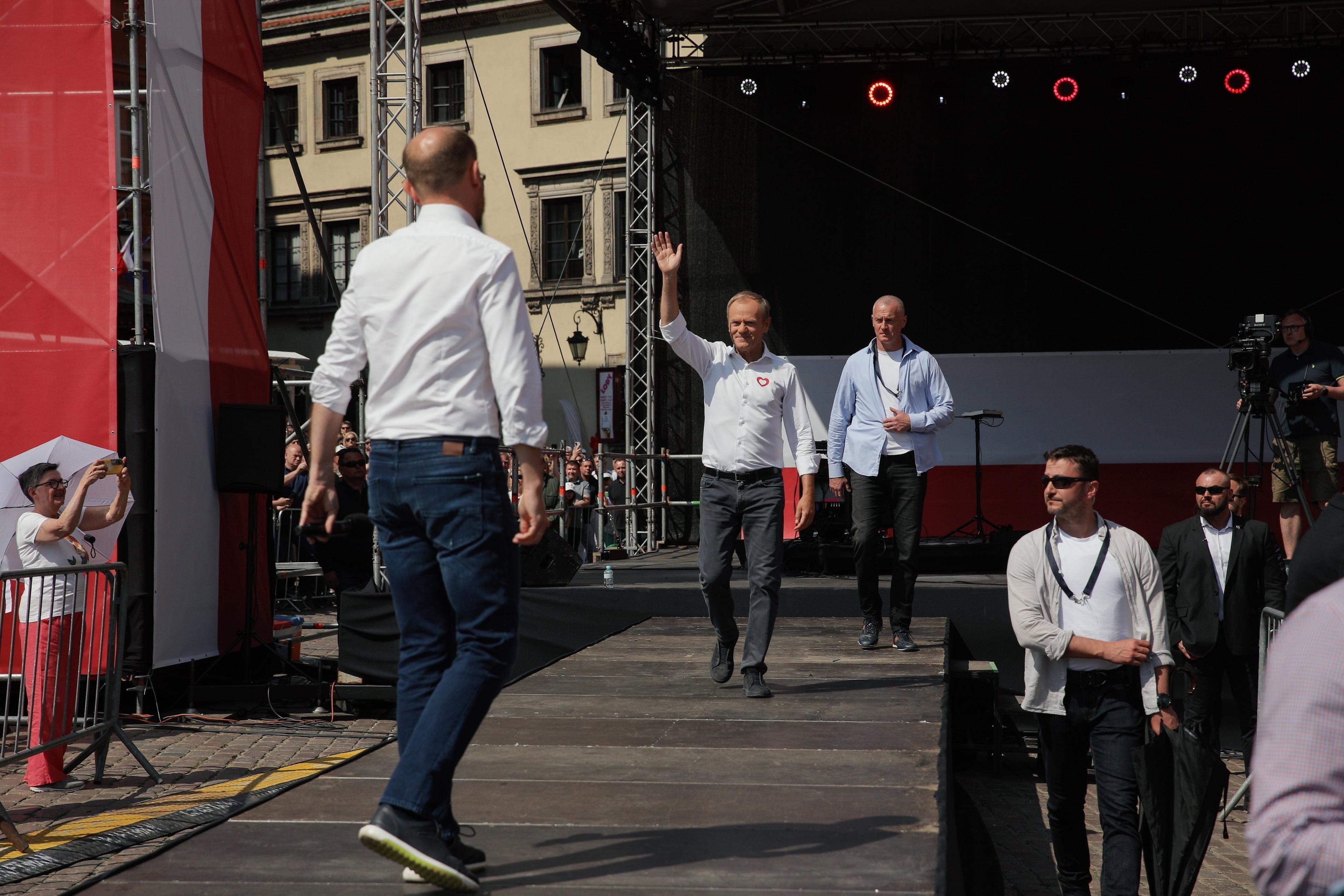 Mężczyźni w białych koszulach na dużej scenie. Pierwszy, Borys Budka, Stoi na pierwszym planie odwrócony plecami do obiektywu, drugi - Donald Tusk - nadchodzi z głębi machając do publiczności