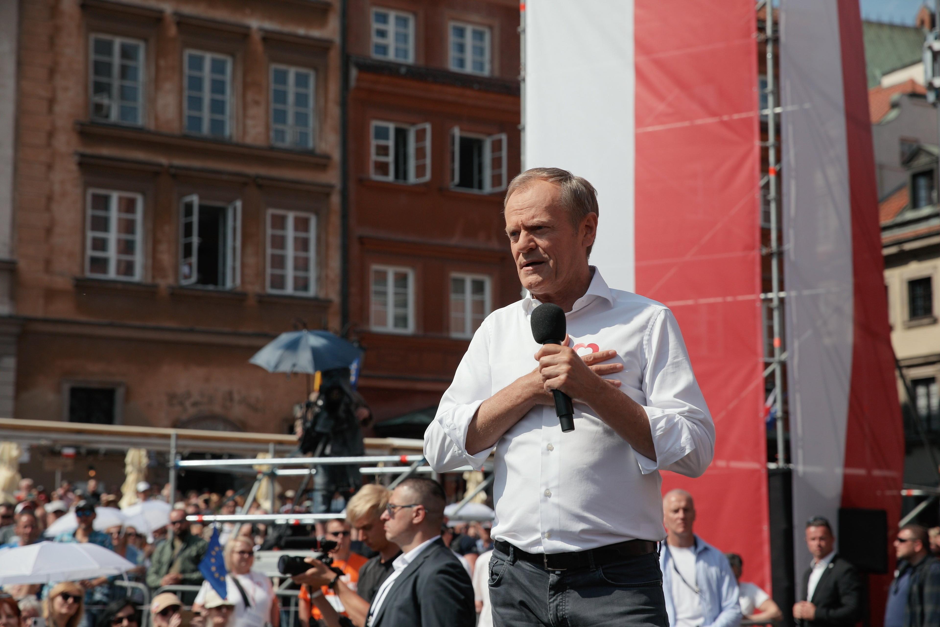 Donald Tusk w białej koszuli z podwiniętymi rękawami. Za nim biało-czerwona flaga