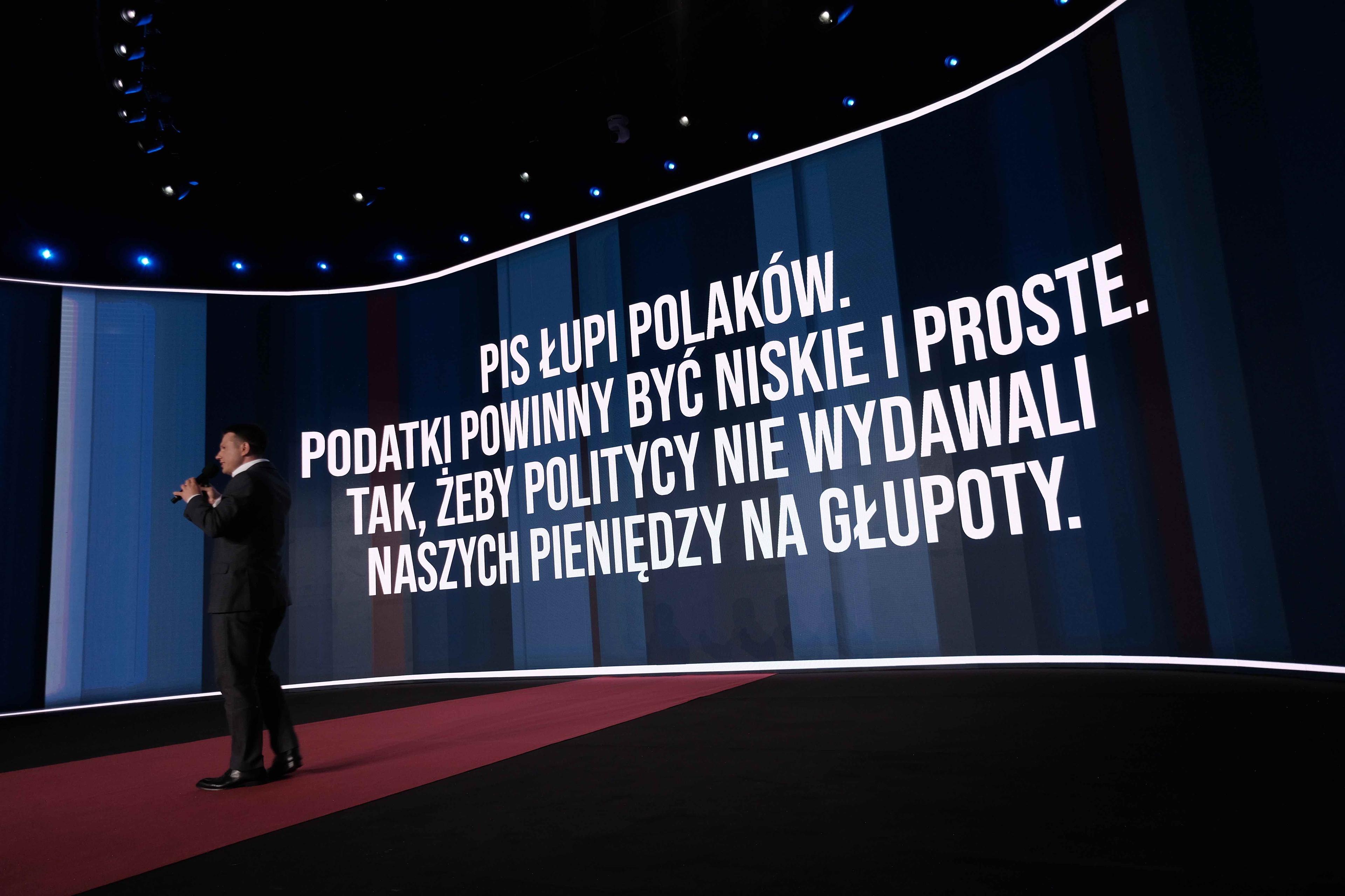 Mężczyzna przemawia na scenie podczas konwencyji partyjnej, w tle na ekranie widać hasło: „PiS łupi Polaków. Podatki powinny być niskie i proste, tak żeby politycy nie wydawali naszych pieniędzy na głupoty.