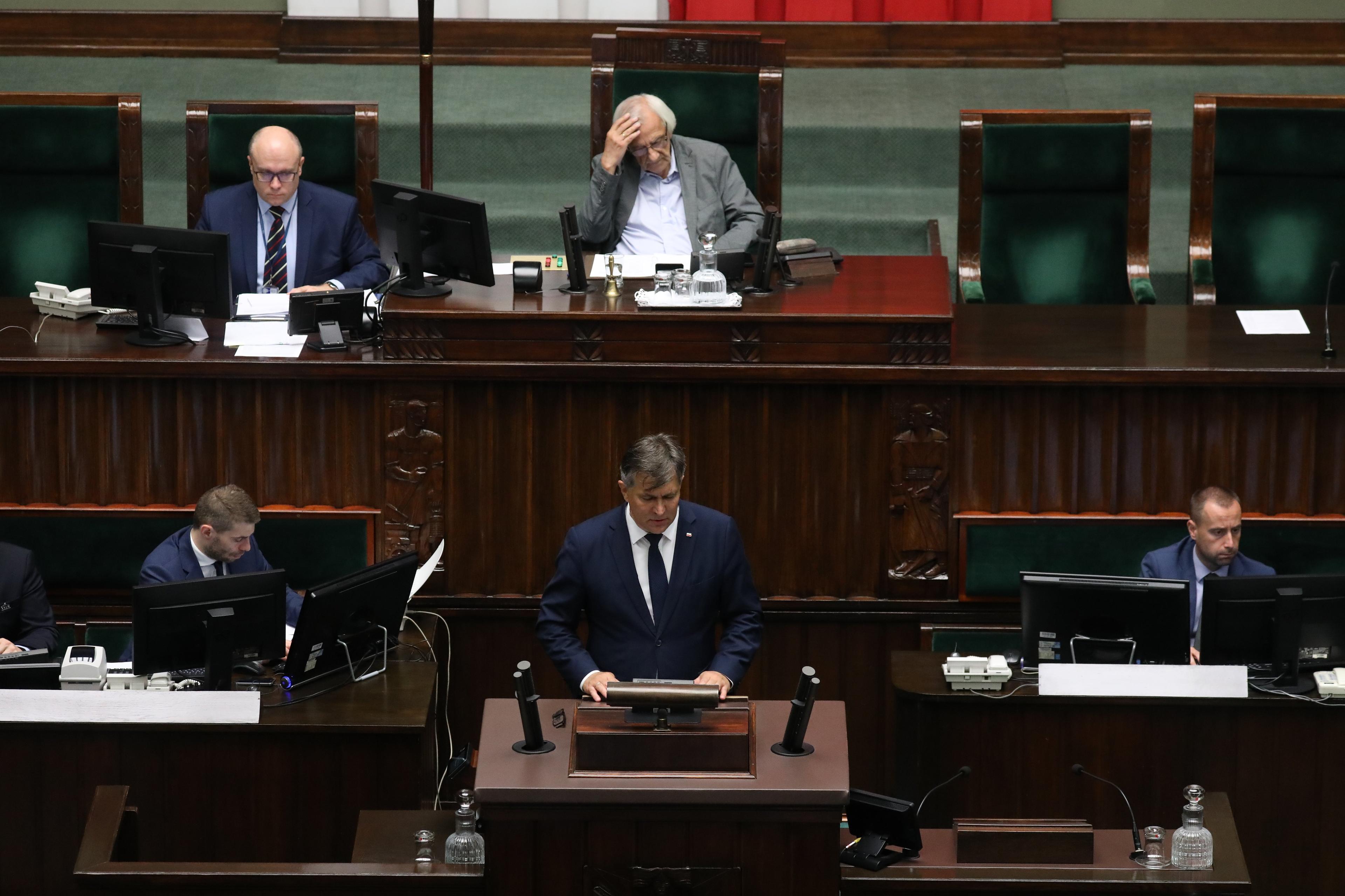 Obrady Sejmu Siwowłosy mężczyzna (prowadzący pbrady wicemarszałek Terlecki z PiS) trzyma się za głowę. Na mównicy męzczyzna w garniturze (poseł)