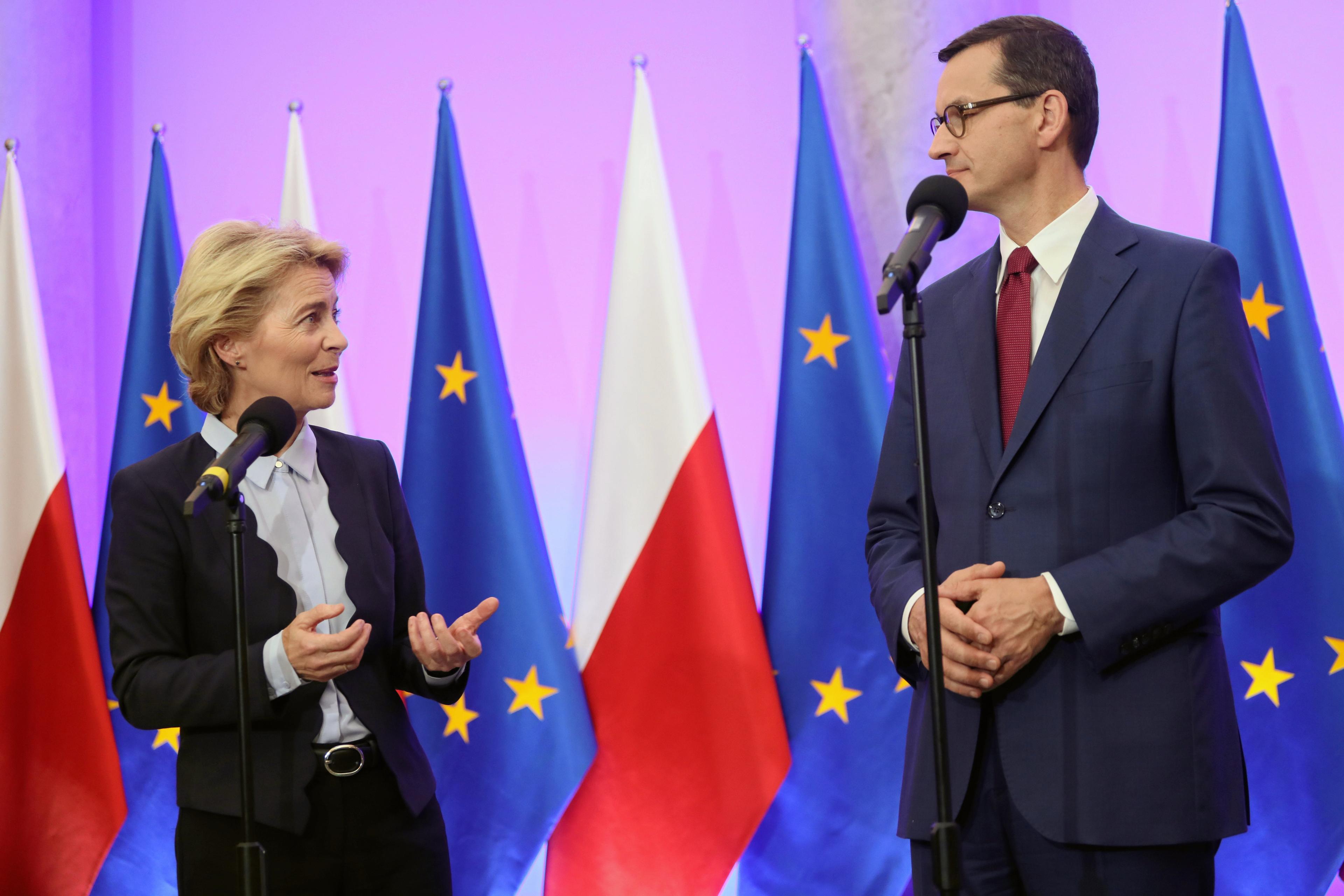 Na zdjęciu widzimy Ursulę von der Leyen i Mateusza Morawieckiego stojących i patrzących się na siebie. Za nimi jest rząd polskich i unijnych flag i różowa ściana.