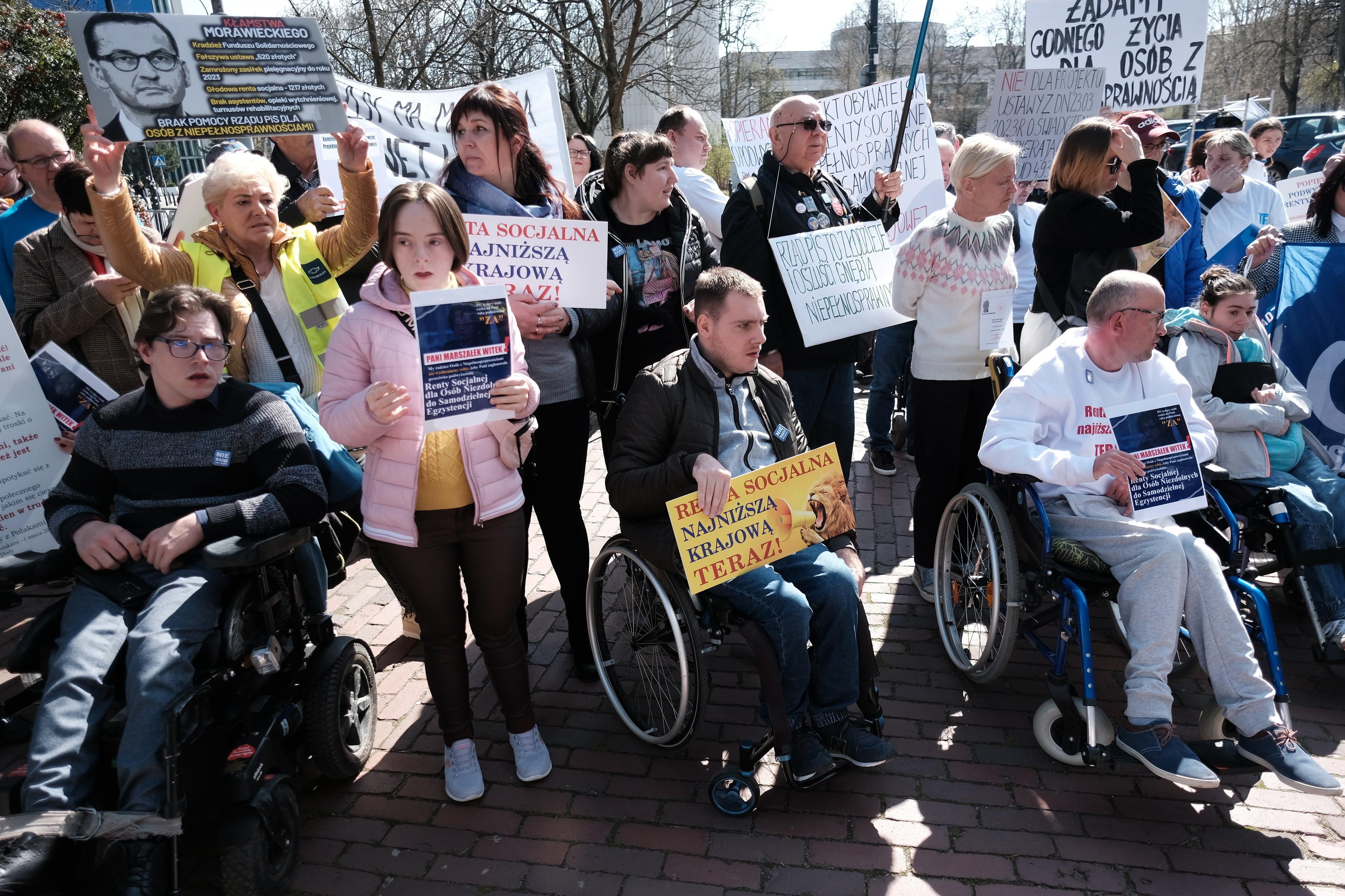 Osoby z niepełnosprawnościami przed Sejmem trzymają w dłoniach banery