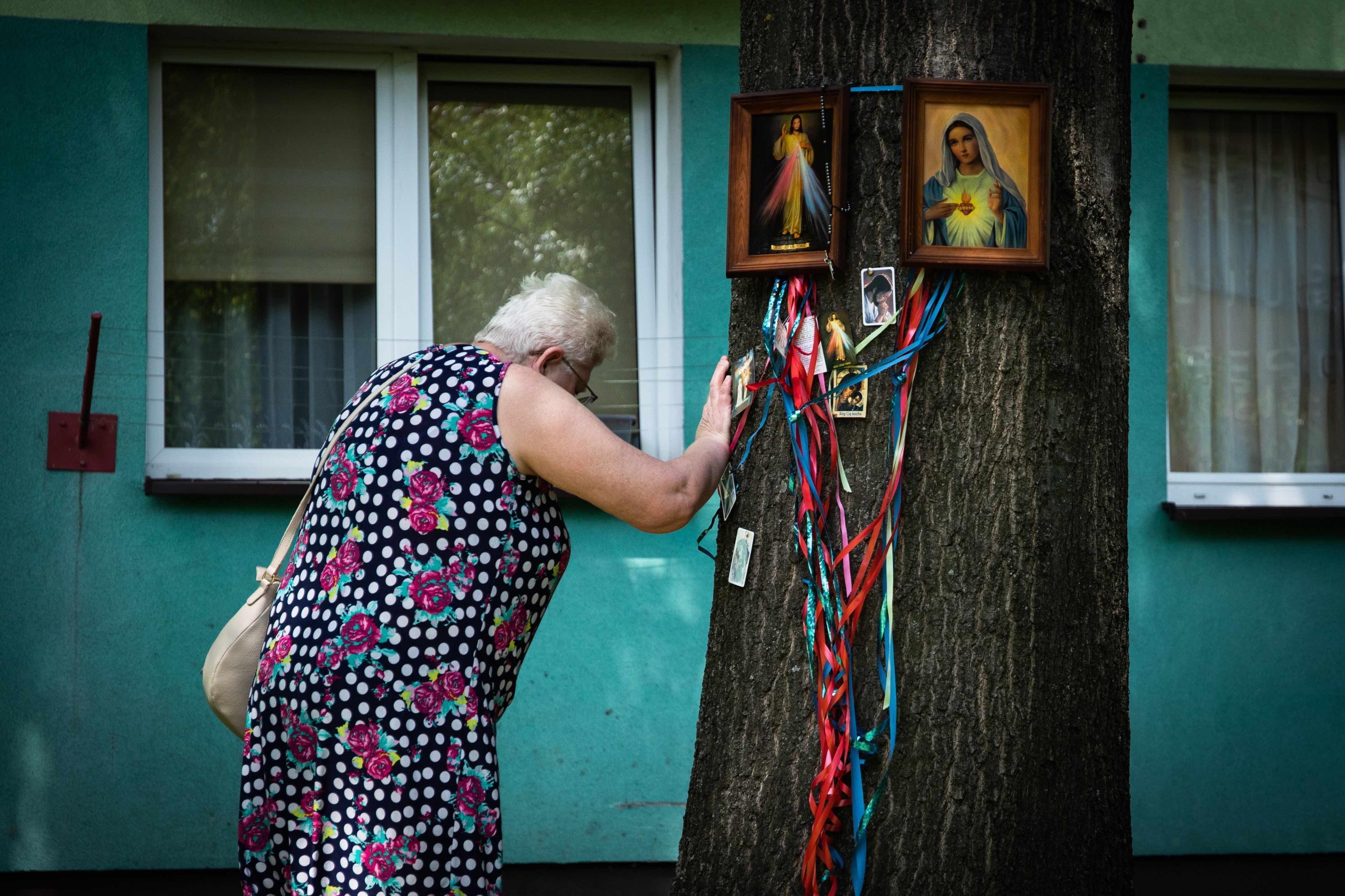 Kobieta opiera rękę o drzewo, na którym wiszą obrazki święte i wstążki. W tle widać okna bloku mieszkalnego.