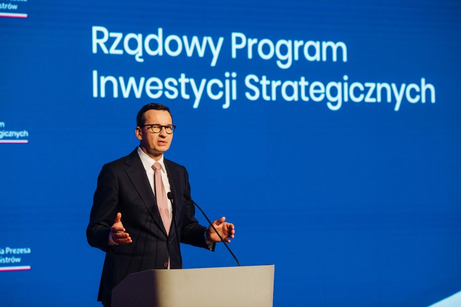 Premier Polski Mateusz Morawiecki w czarnym garniturze stoi na scenie Makowskiego Domu Kultury na tle dużego niebieskiego ekranu z napisem Rządowy Program Inwestycji Strategicznych