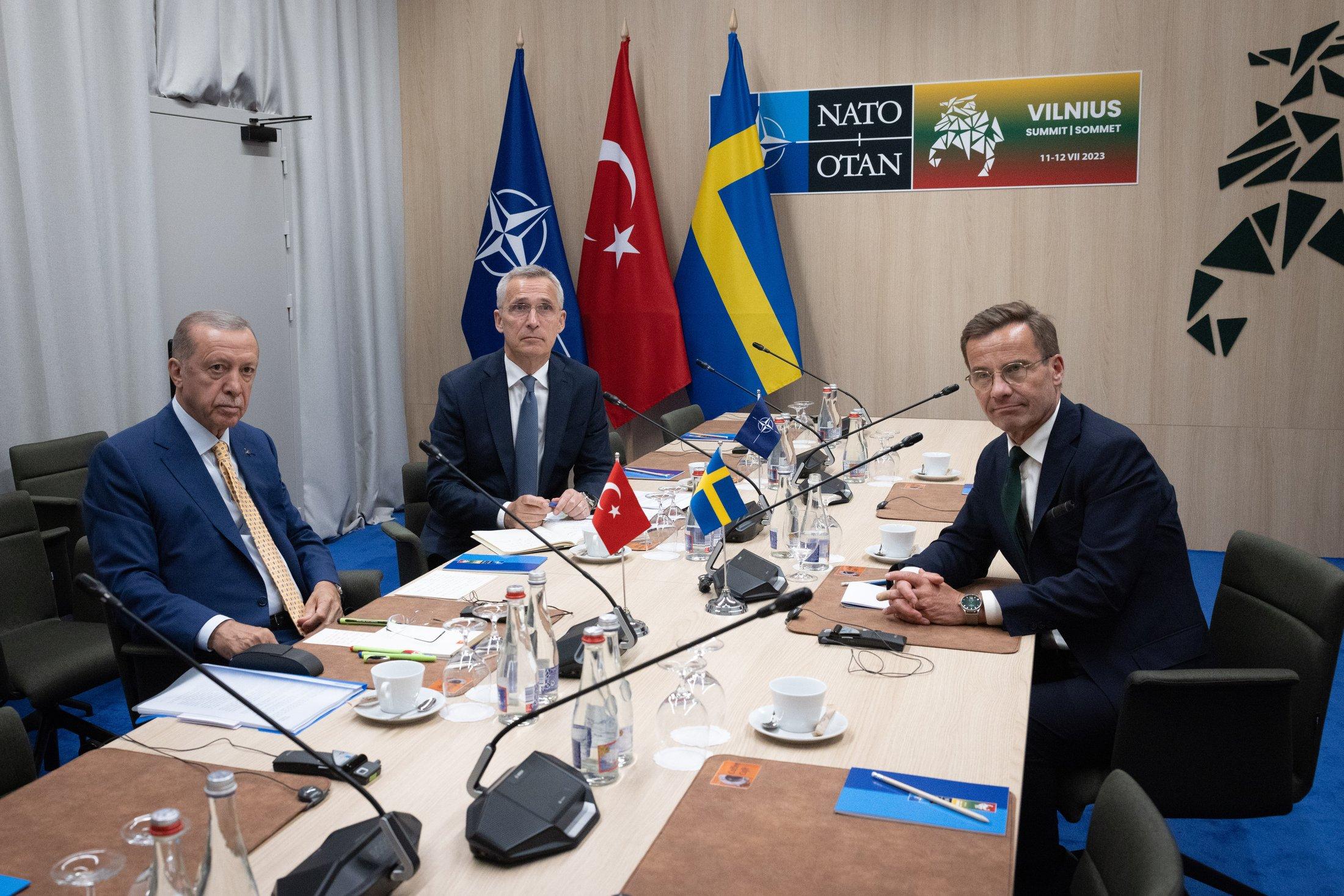 Trzech mężczyzn siedzi przy stole negocjacyjnym, w rogu pomieszczania trzy flagi: flaga NATO, Turcji i Szwecji. Mężczyźni na zdjęciu to od lewej prezydent Turcji Recep Tayyip Erdogan, w środku sekretarz generalny NATO Jens Stoltenberg oraz po przeciwnej stronie stołu premier Szwecji, Ulf Kristersson. Mężczyźni patrzą w obiektyw aparatu, przed nimi trudne rozmowy o odblokowaniu przez Turcję członkostwa Szwecji w Sojuszu.
