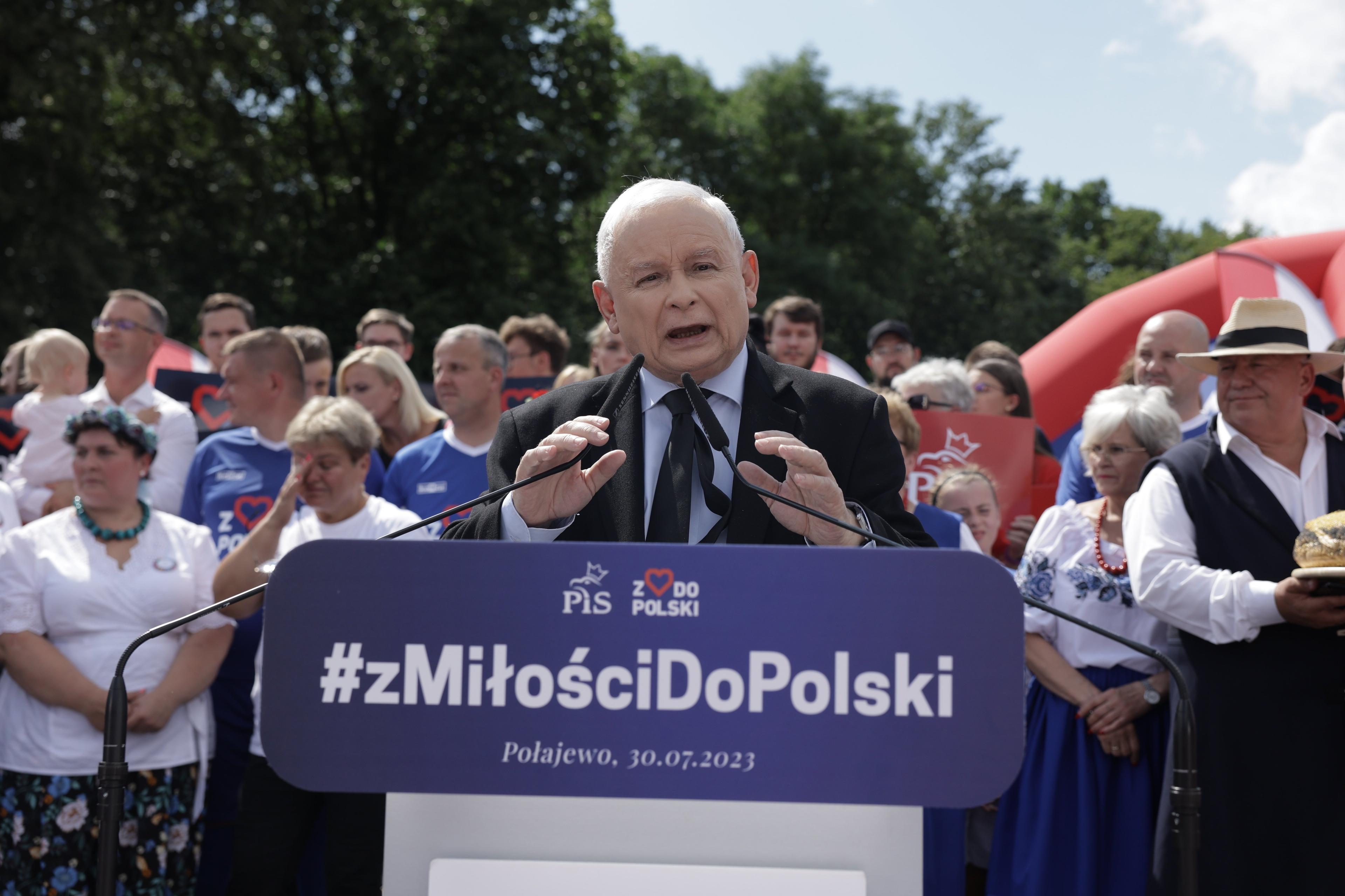 siwy mężczyzna przemawia na tle drzew i zebranych na spotkaniu ludzi, stoi przed pulpitem z napisem z Miłości do Polski