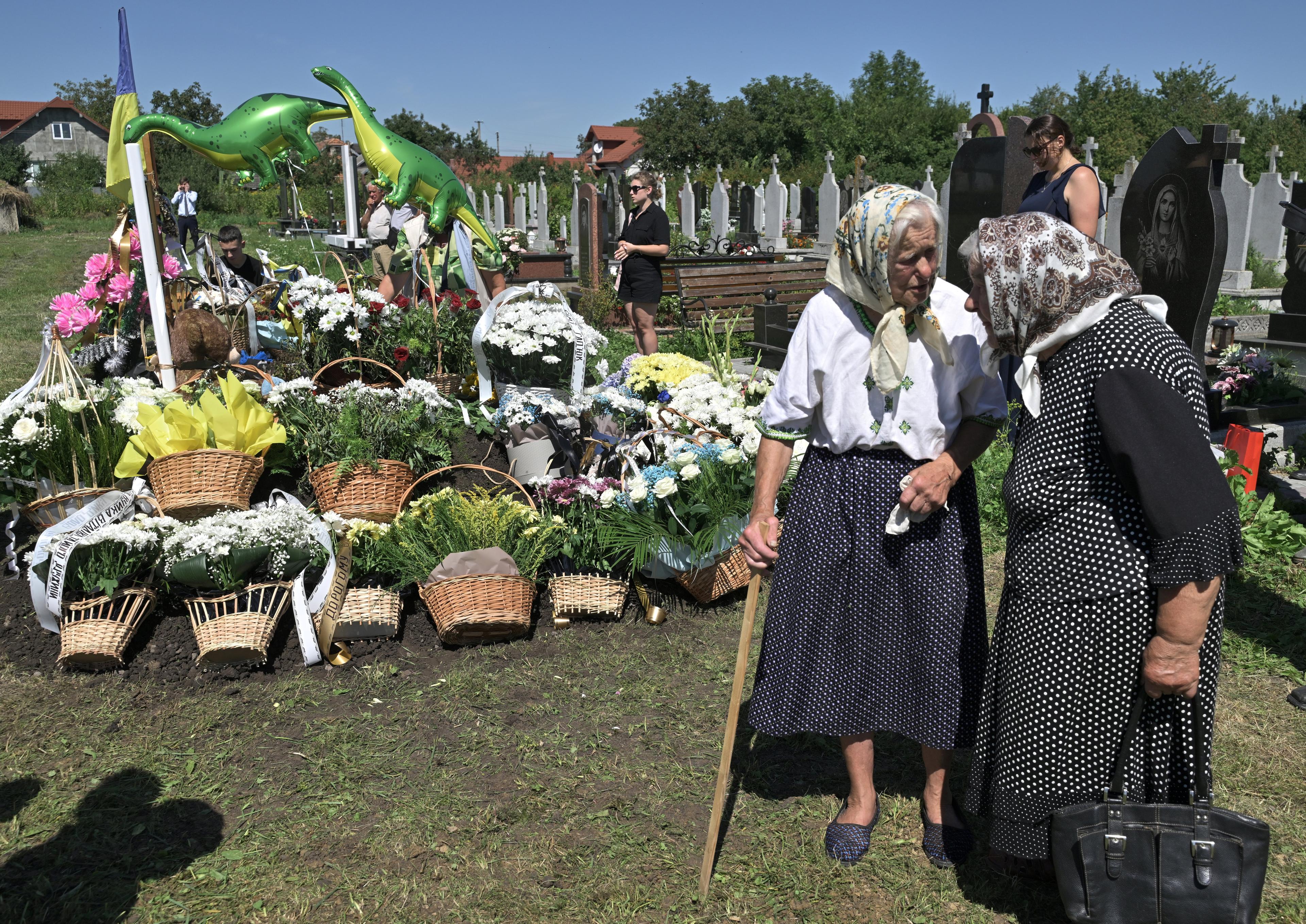 dwie staruszki w chustkach przed świeżym grobem udekorowanym koszykami z kwiatami