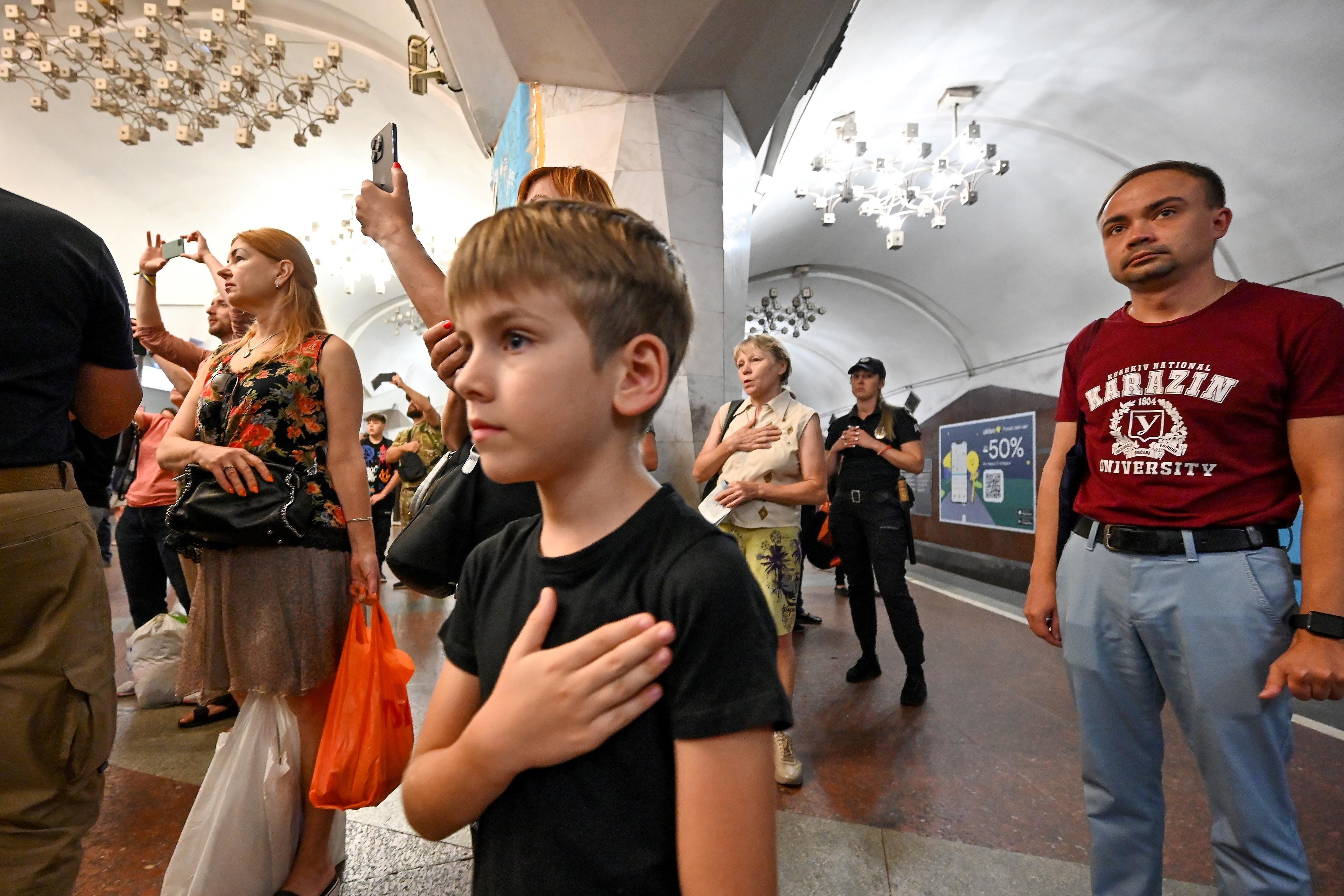 Ludzie na peronie w metrze śpiewają trzymając prawą rękę na sercu. Na pierwszym planie - mały chłopiec