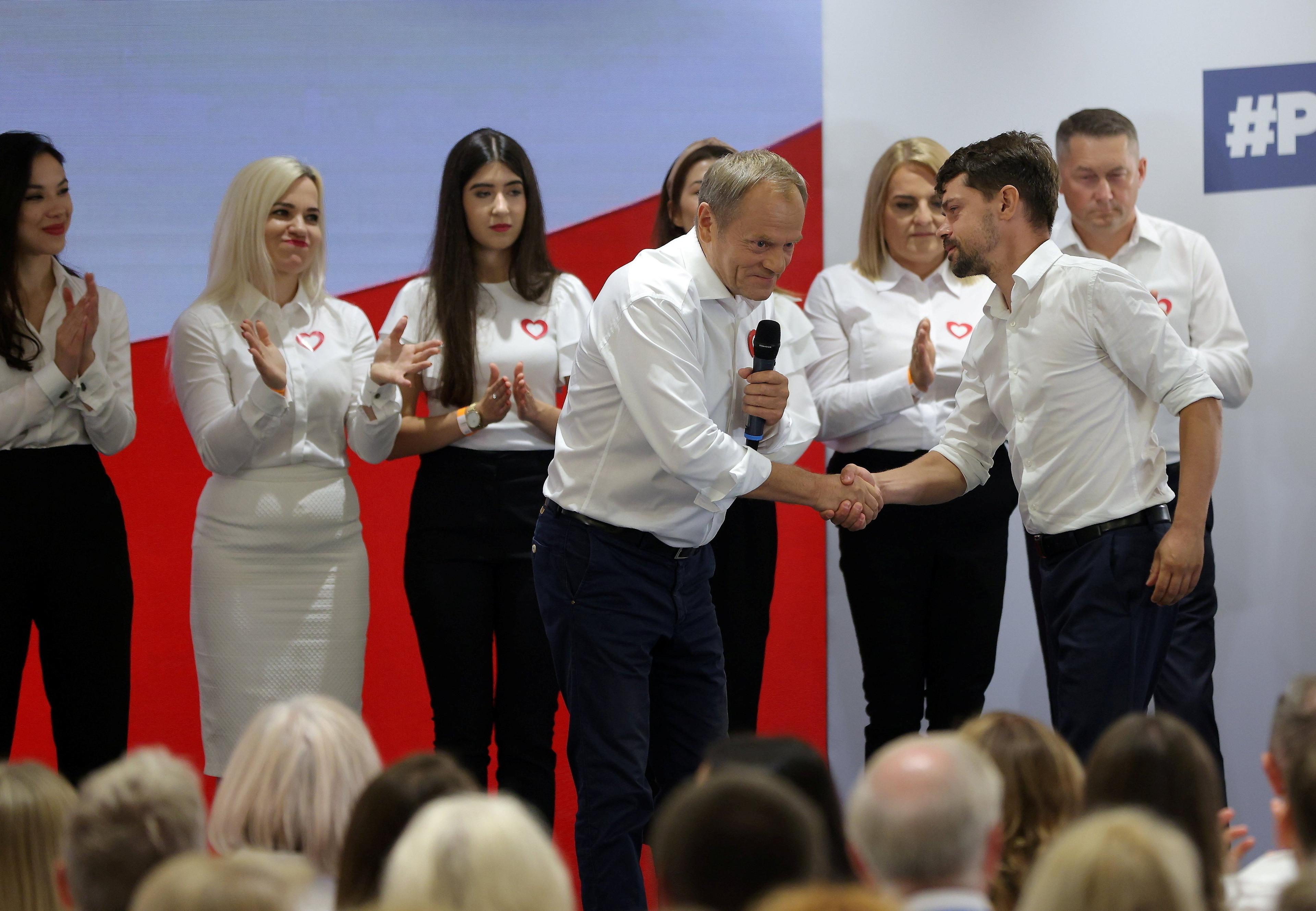 Dwóch mężczyzn - Donald Tusk i Michał Kołodziejczak - w białych koszulach z podwiniętymi rękawami, podaje sobie ręce na scenie