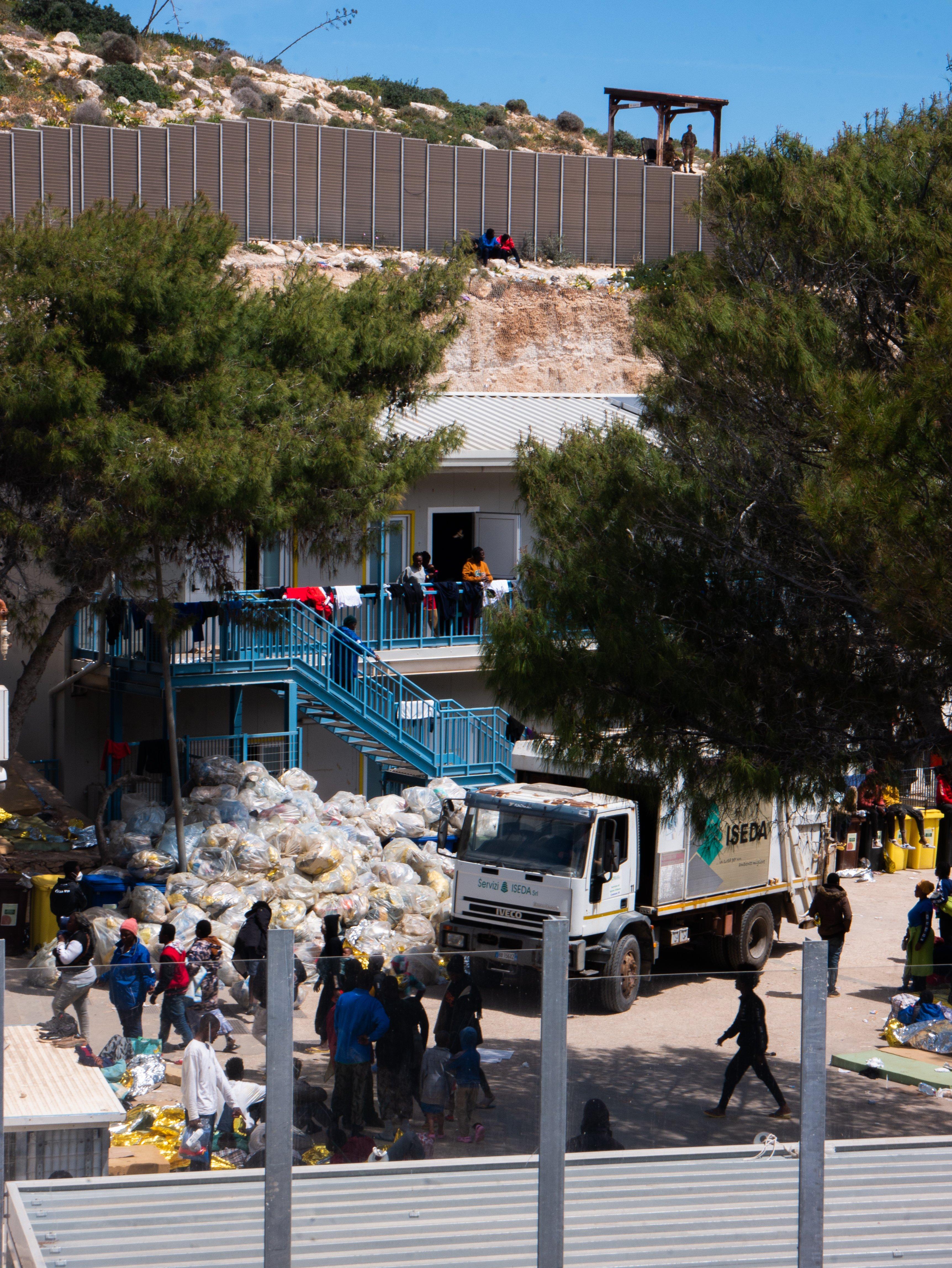 Ogrodzony teren ośrodka dla uchodźców, ludzie noszą worki ze śmieciami