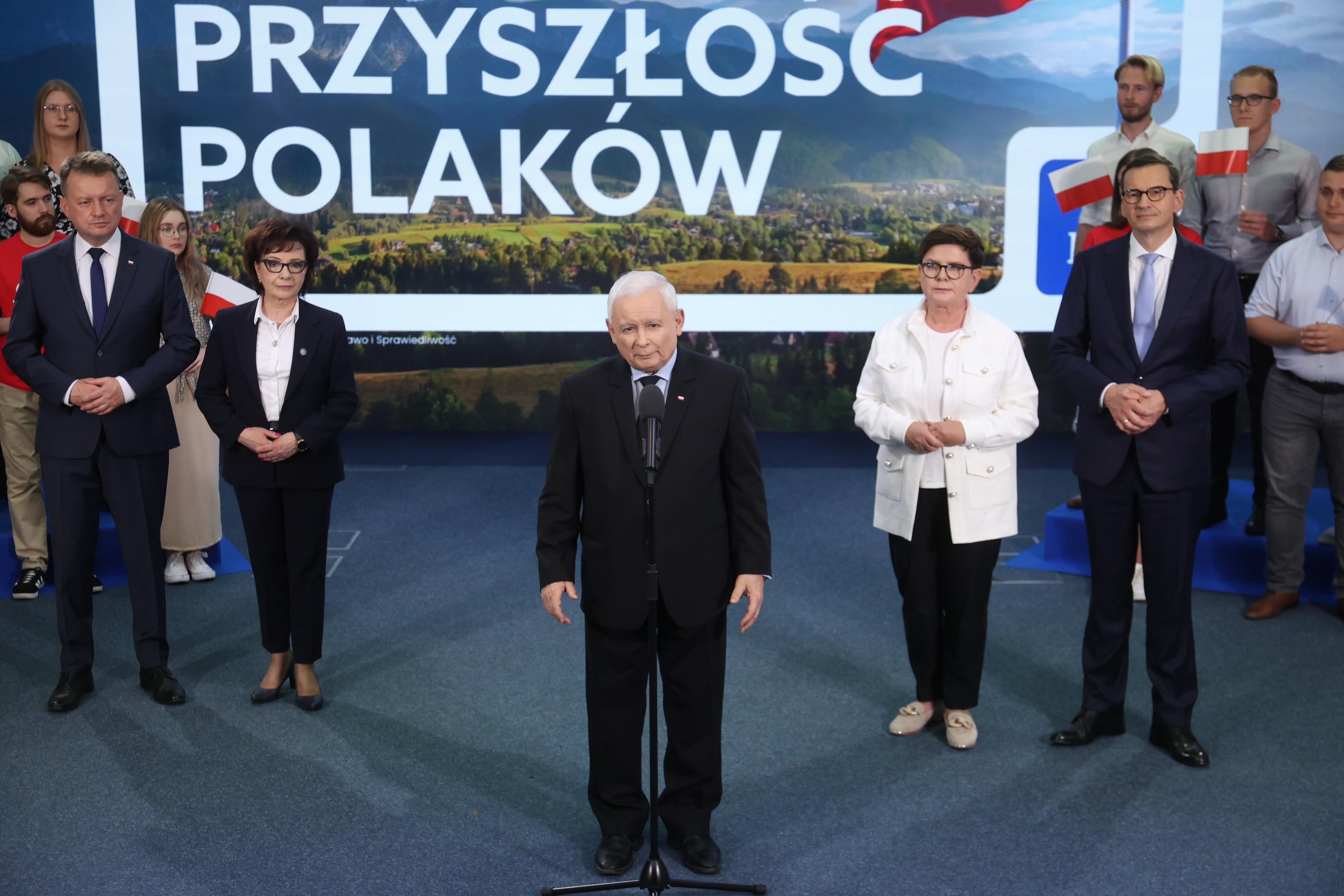 Mężczyzna, starszy, siwy, niski, Jarosław Kaczyński , stoi na scenie na tle hasła wyborczego PiS "Bezpieczna przyszłość Polaków'