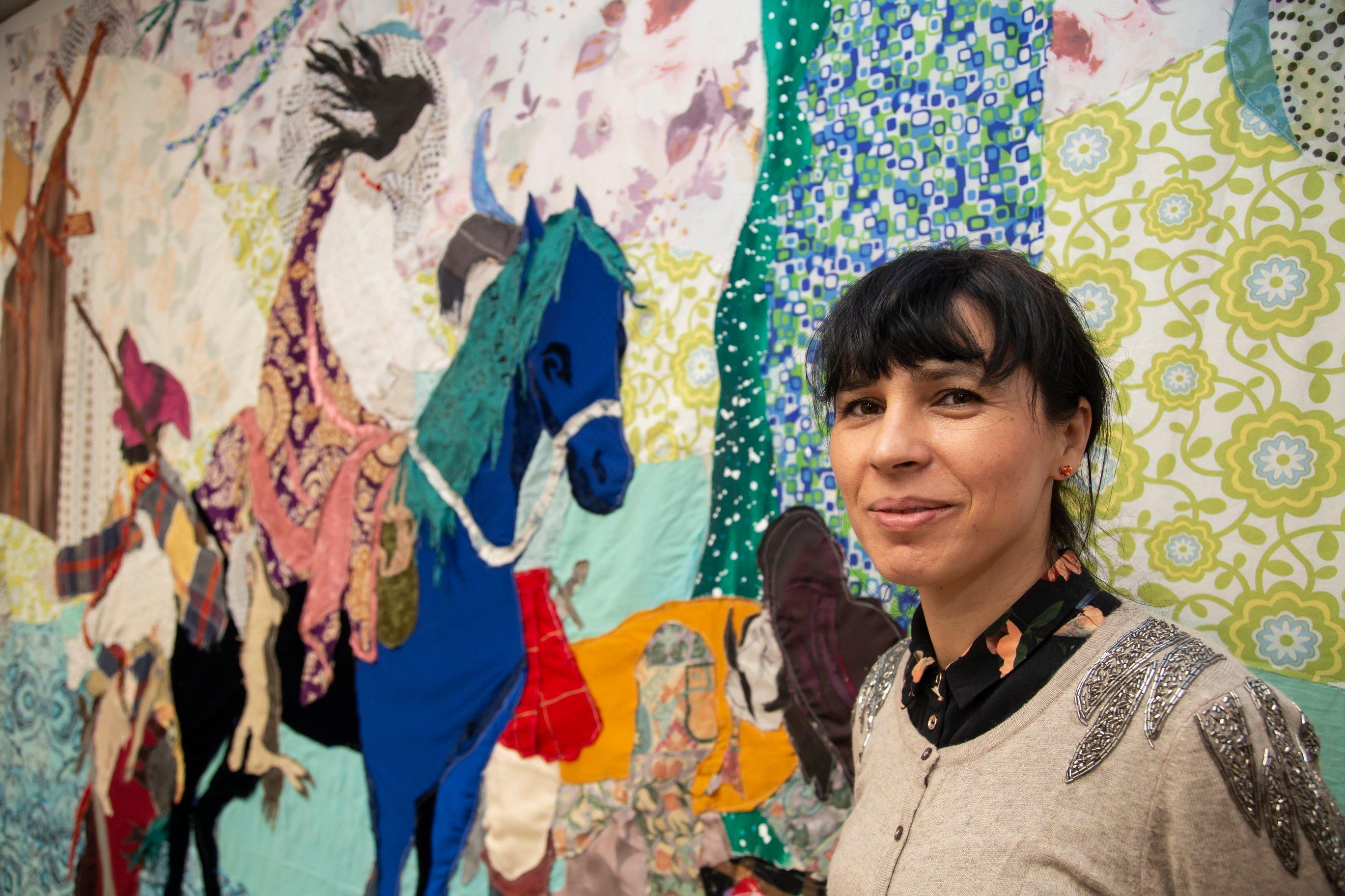 Artystka Małgorzata Mirga-Tas stoi na tle swojej pracy, przedstawiającej jeźdźca na koniu