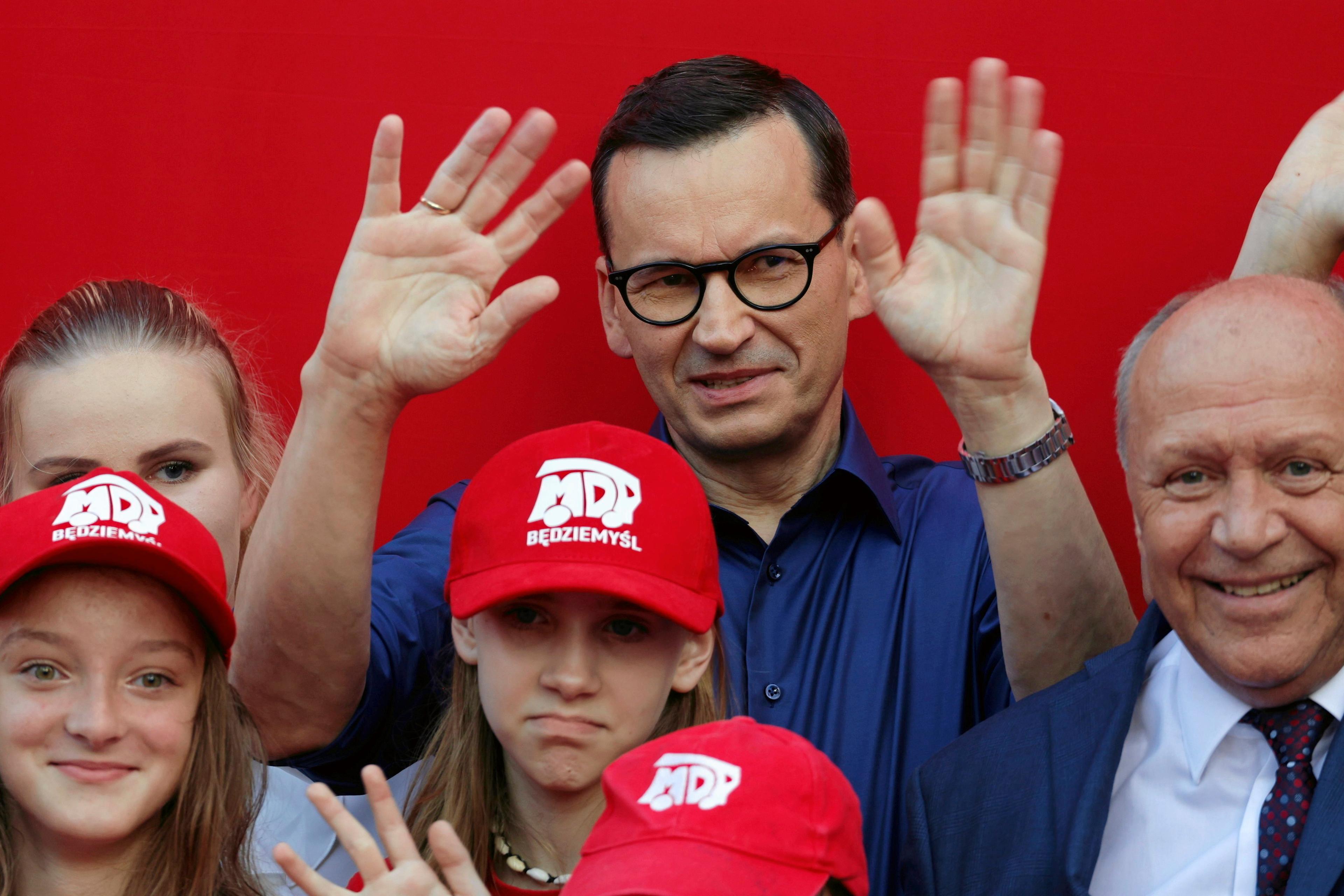Premier Mateusz Morawiecki w niebieskiej koszuli macha do kamery na tle czerwonej ściany razem z dziećmi i innym mężczyzną