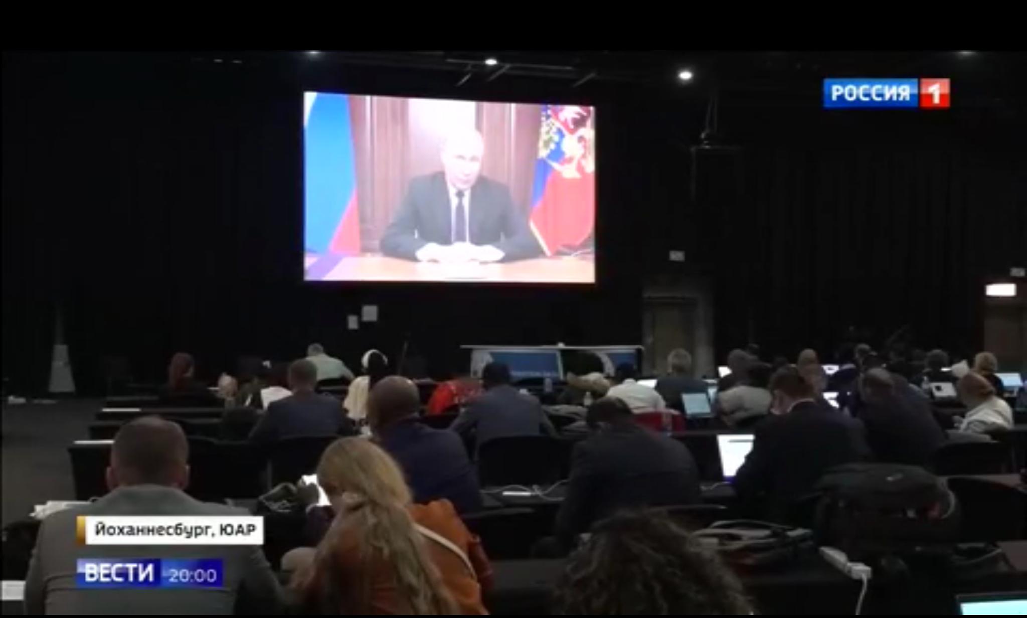 Sala konferencyjna. Na ekranie przemawiający Putin