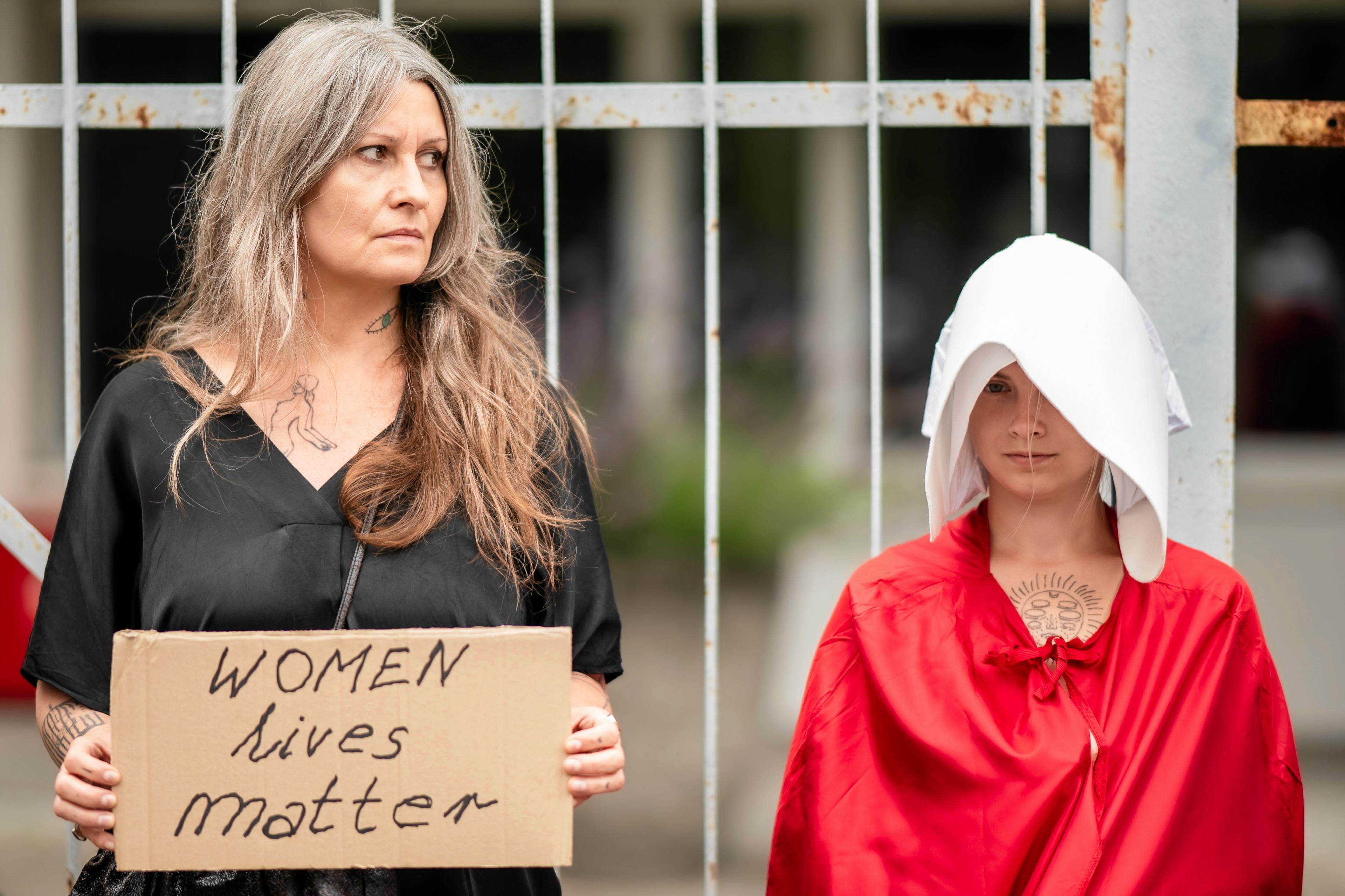 Kobieta stoi z transparentem "woman lives matter". A obok niej druga kobieta przebrana za "podręczną"