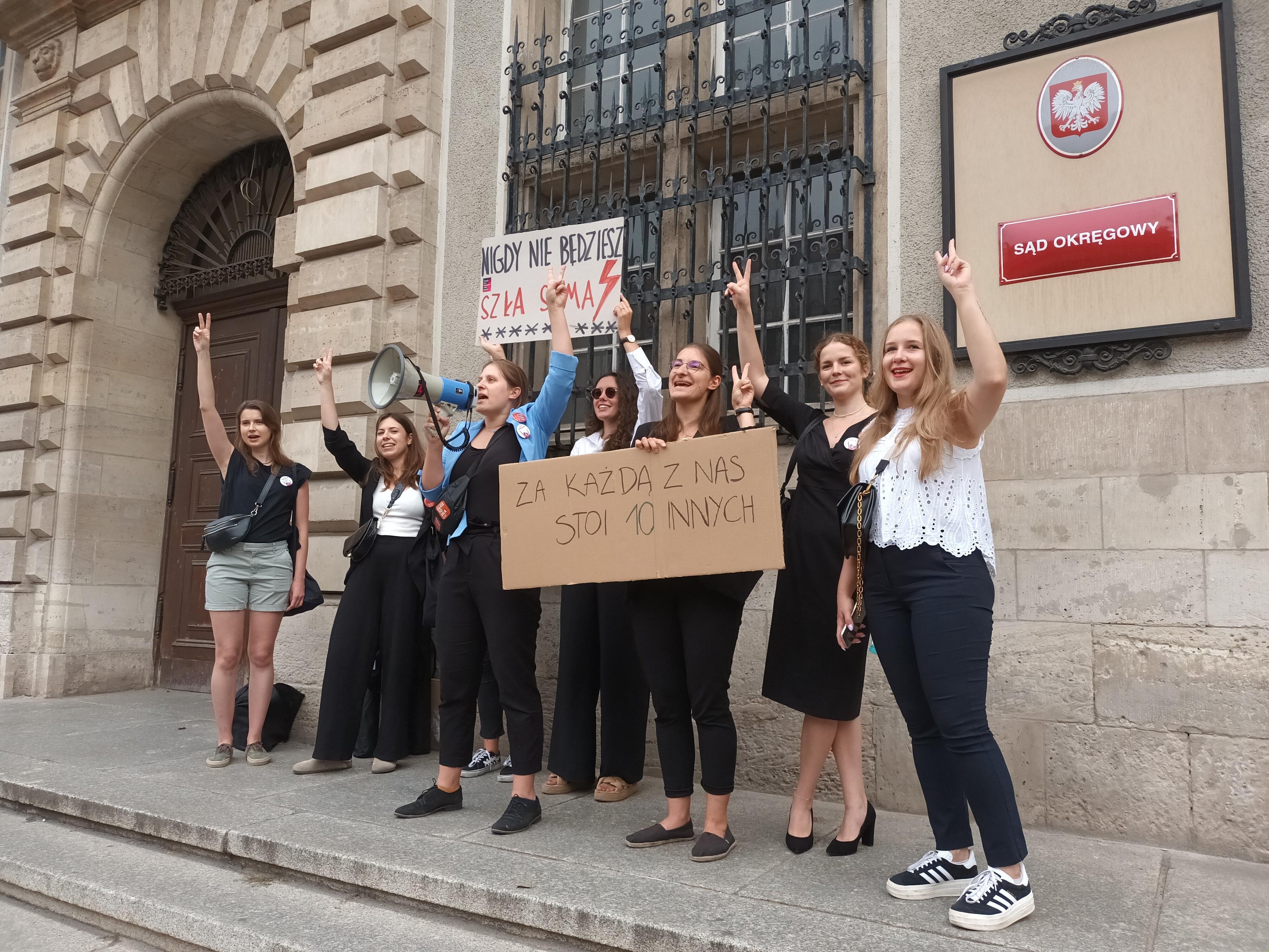 Kobiety przed sądem okręgowym. Trzymają transparenty "Nigdy nie będziesz szła sama" i "Za każdą z nas stoi 10 innych". Wśród kobuet - młoda osoba z długimi włosami w ciemnej sukience- Julia Landowska