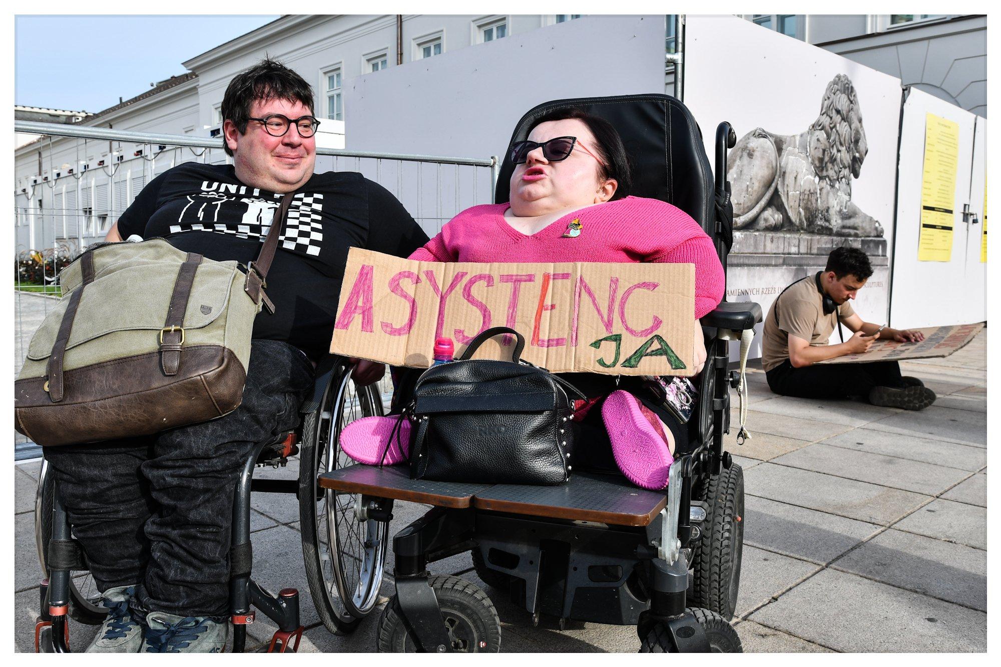 Dwie osoby z niepełnosprawnością na wózkach przed pałacem prezydenckim. Jedna trzyma transparent z napisem "asystenc-ja"
