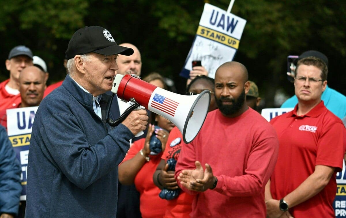 Prezydent Biden z czapce z daszkiem z megofonem w ręku, w tle klaszcząa mu strajkujący