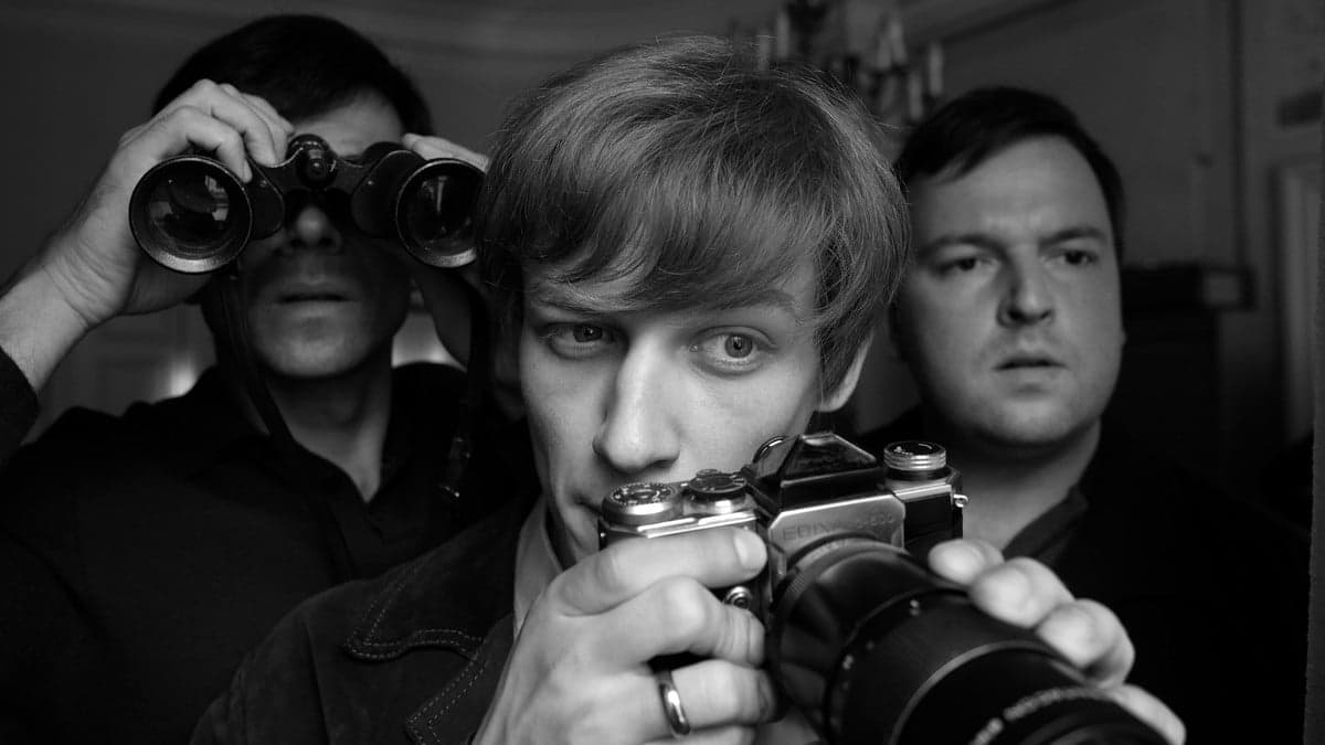 Mężczyzna z aparatem fotograficznym, w tle mężczyzna patrzący przez lornetkę