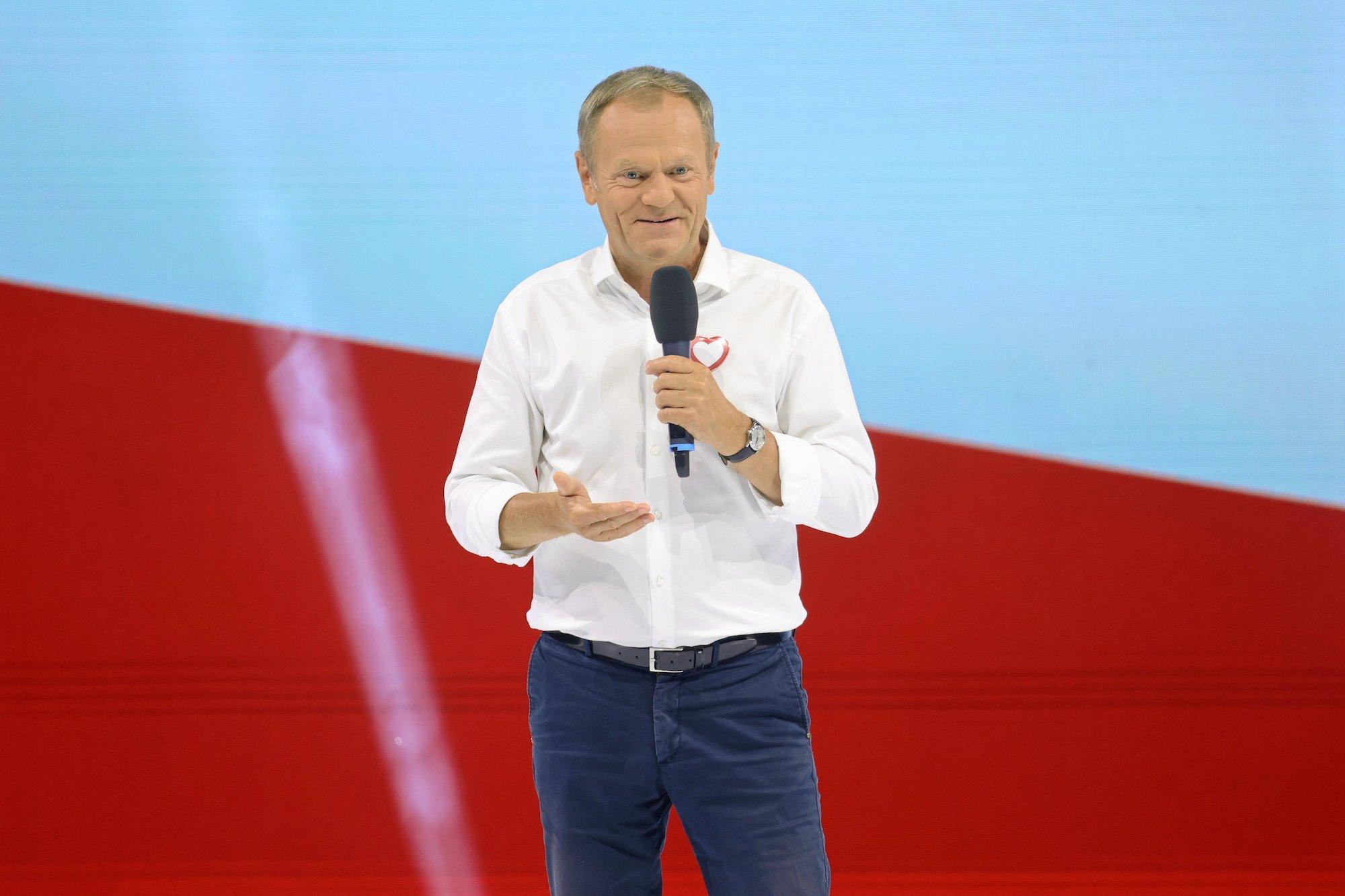 Donald Tusk na konwencji PO w Tarnowie. Przemawia na tle biało czerwonej flagi ubrany w białą koszulę i granatowe spodnie.