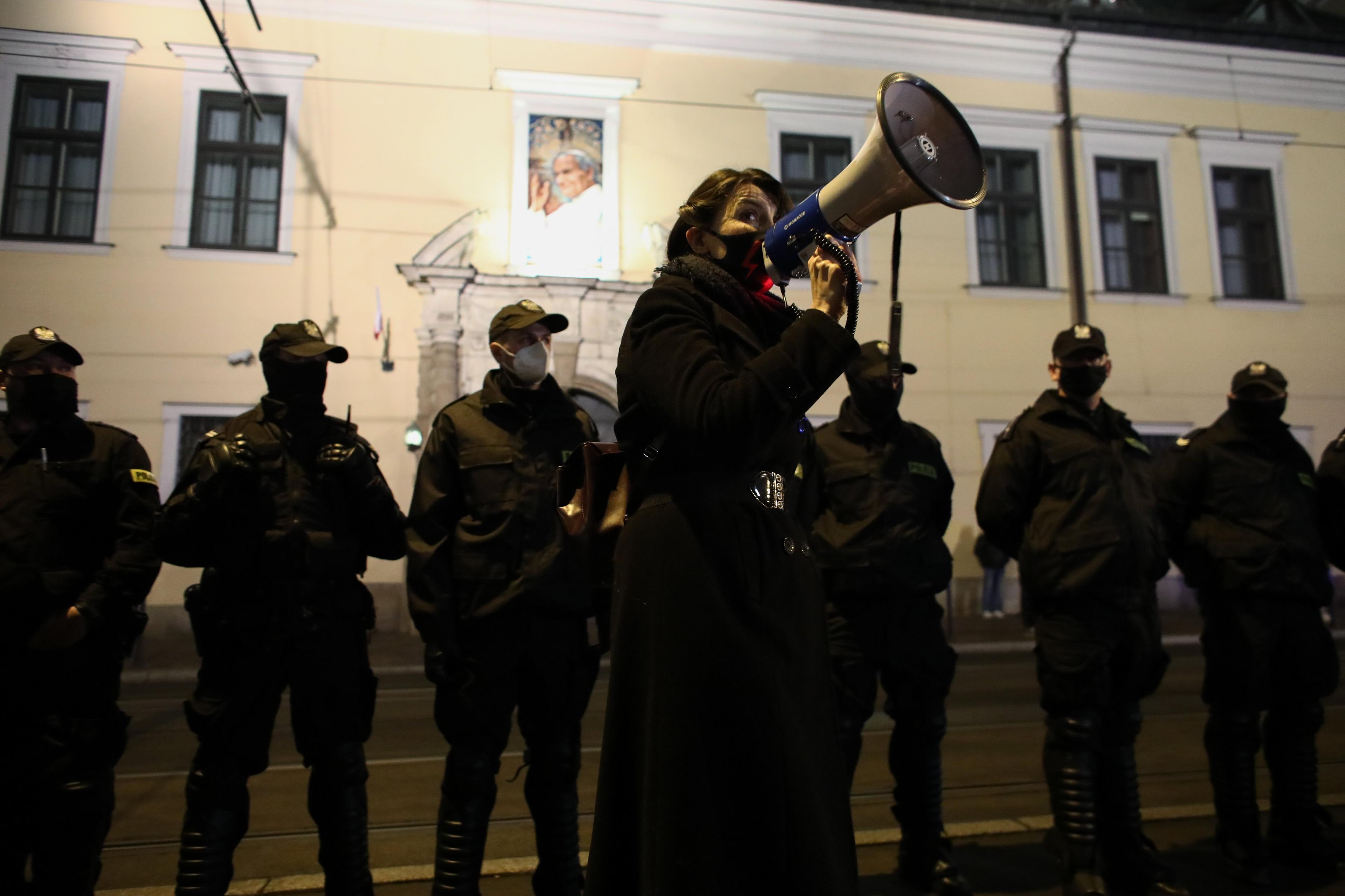 Krakowska kuria z portretem Jana Pawła II otoczona przz policje. Na pierwszym planie protetsująca kobieta z megafonem