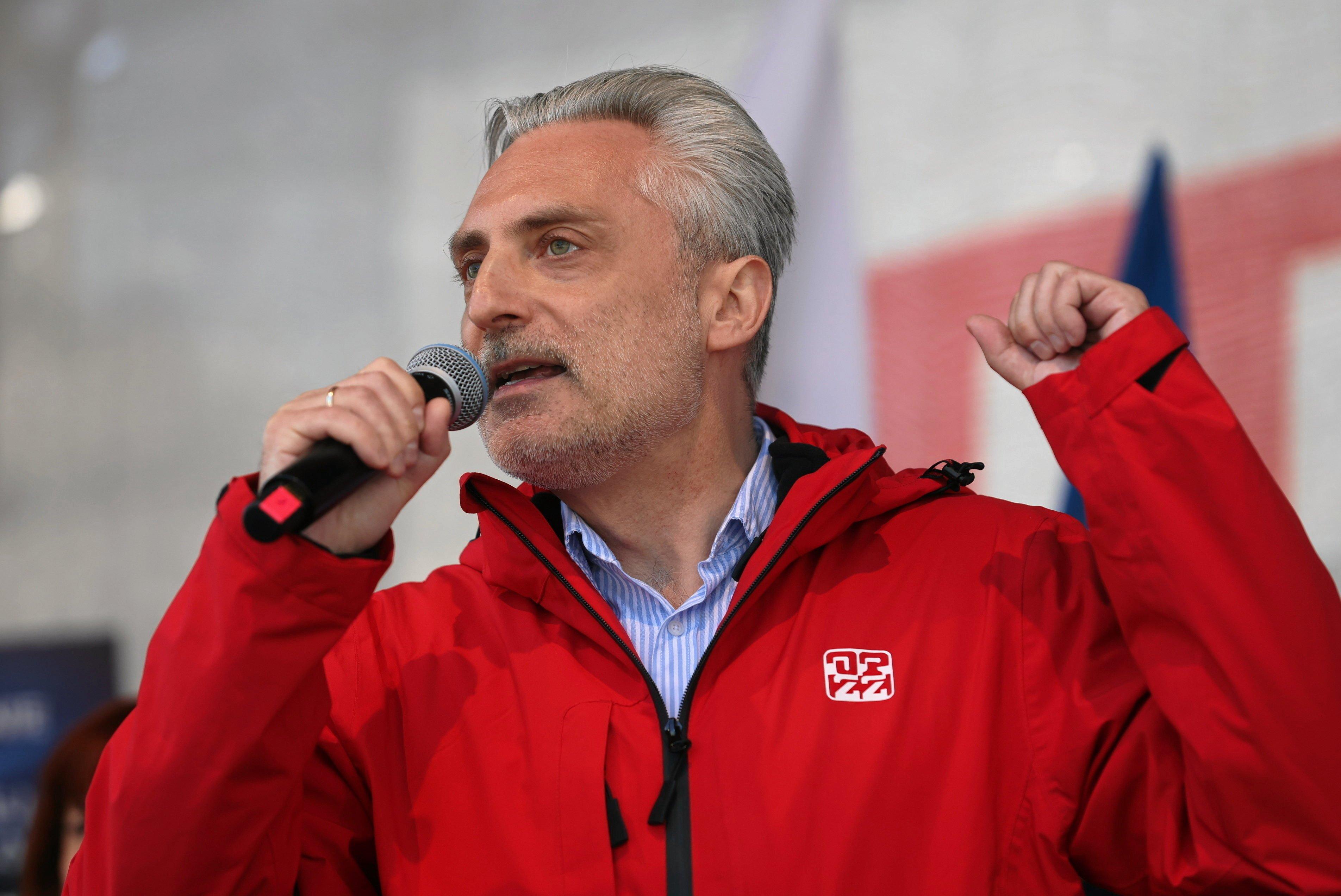 Mężczyzna w siwych włosach przemawia do mikrofonu z podniesionymi rękami, nosi czerwoną kurtkę