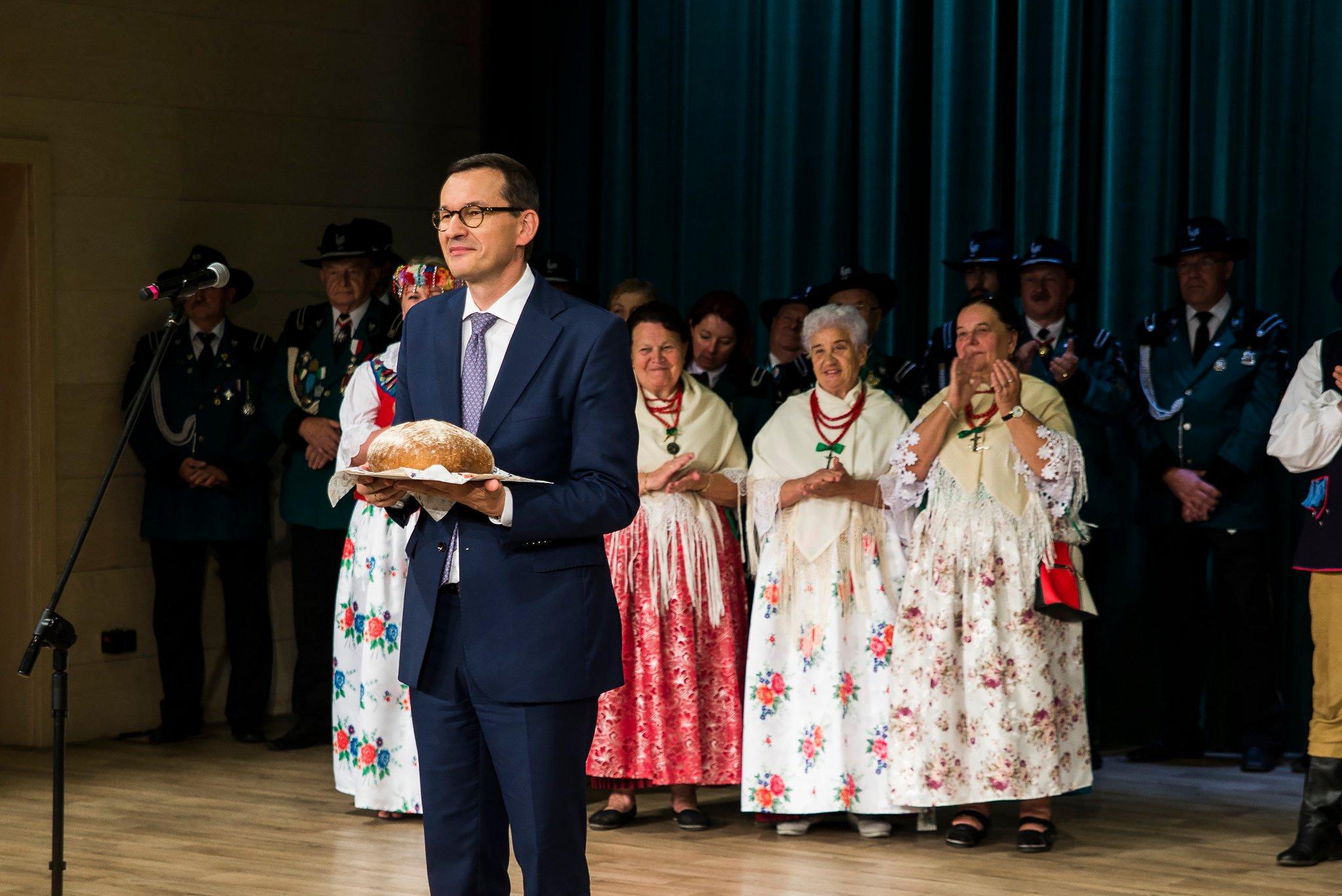 premier Morawiecki stoi na scenie z chlebem w ręce, w tle kobiety w starszym wieku, w strojach ludowych