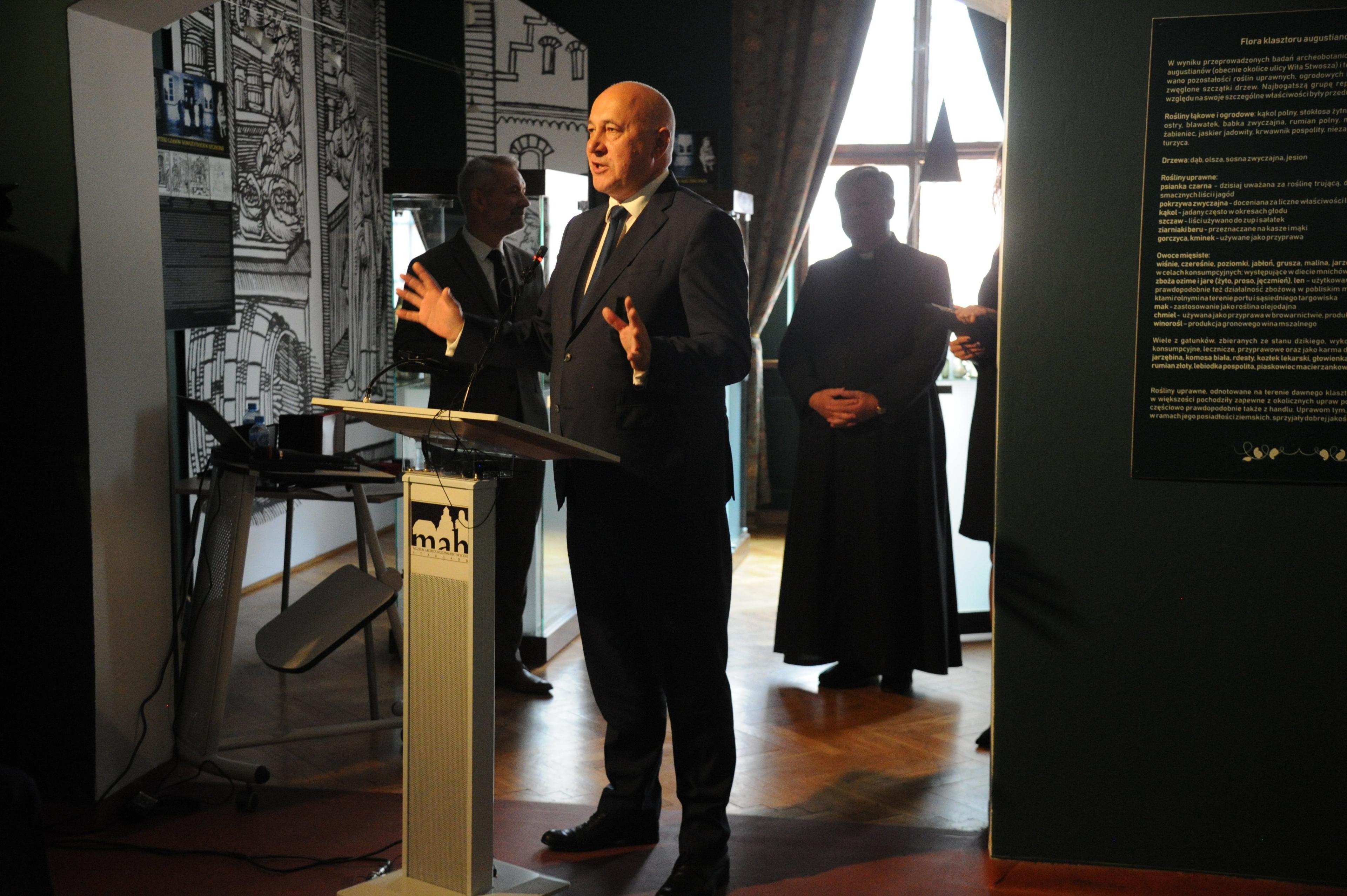 Męzczyzna (Joachim Brudziński) przemawia w ciemnym, przypominającym muzeum pomiszczeniu, z tyłu sylwetka księdza