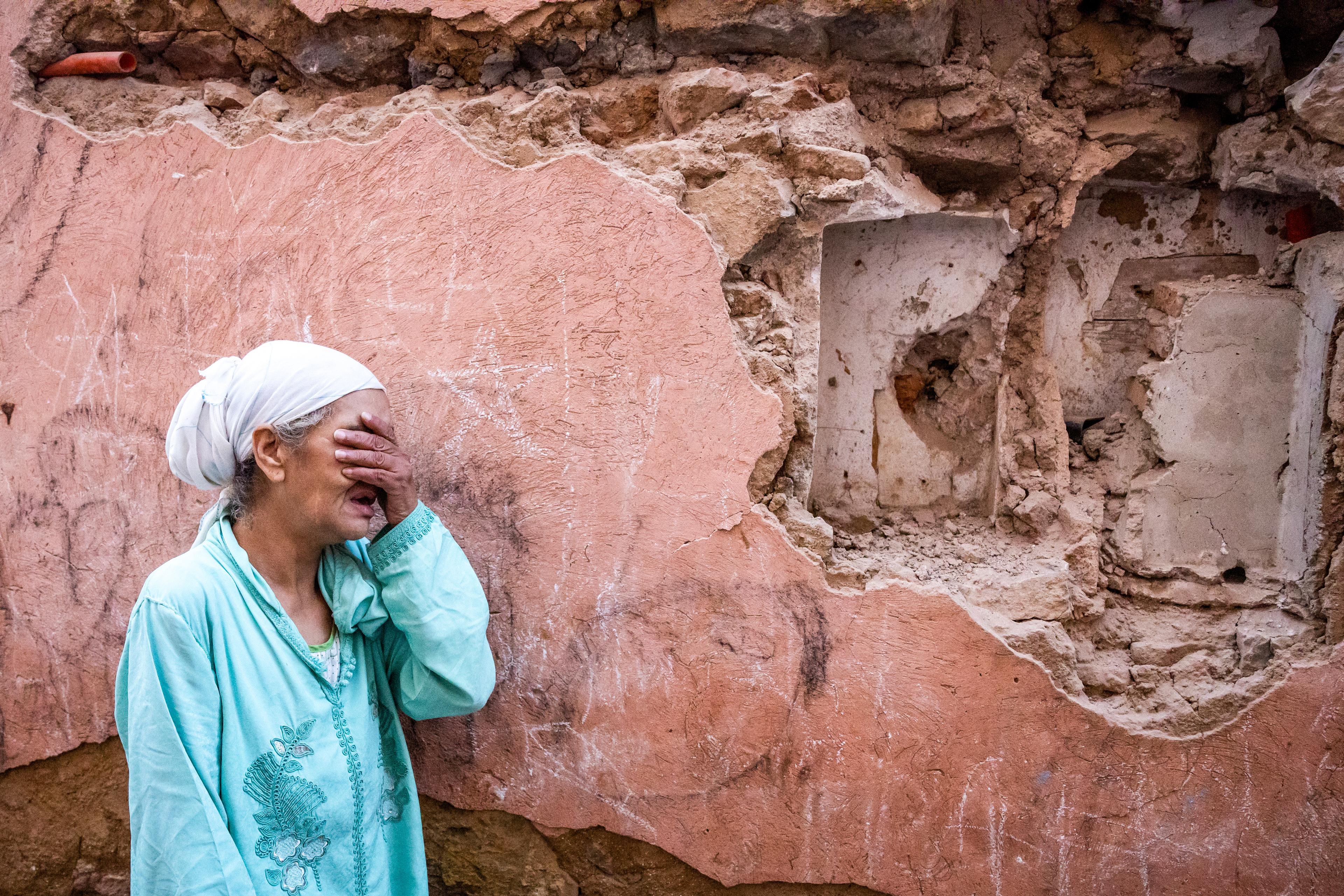 Kobieta płacząca zasłania oczy, w tle zrujnowany budynek