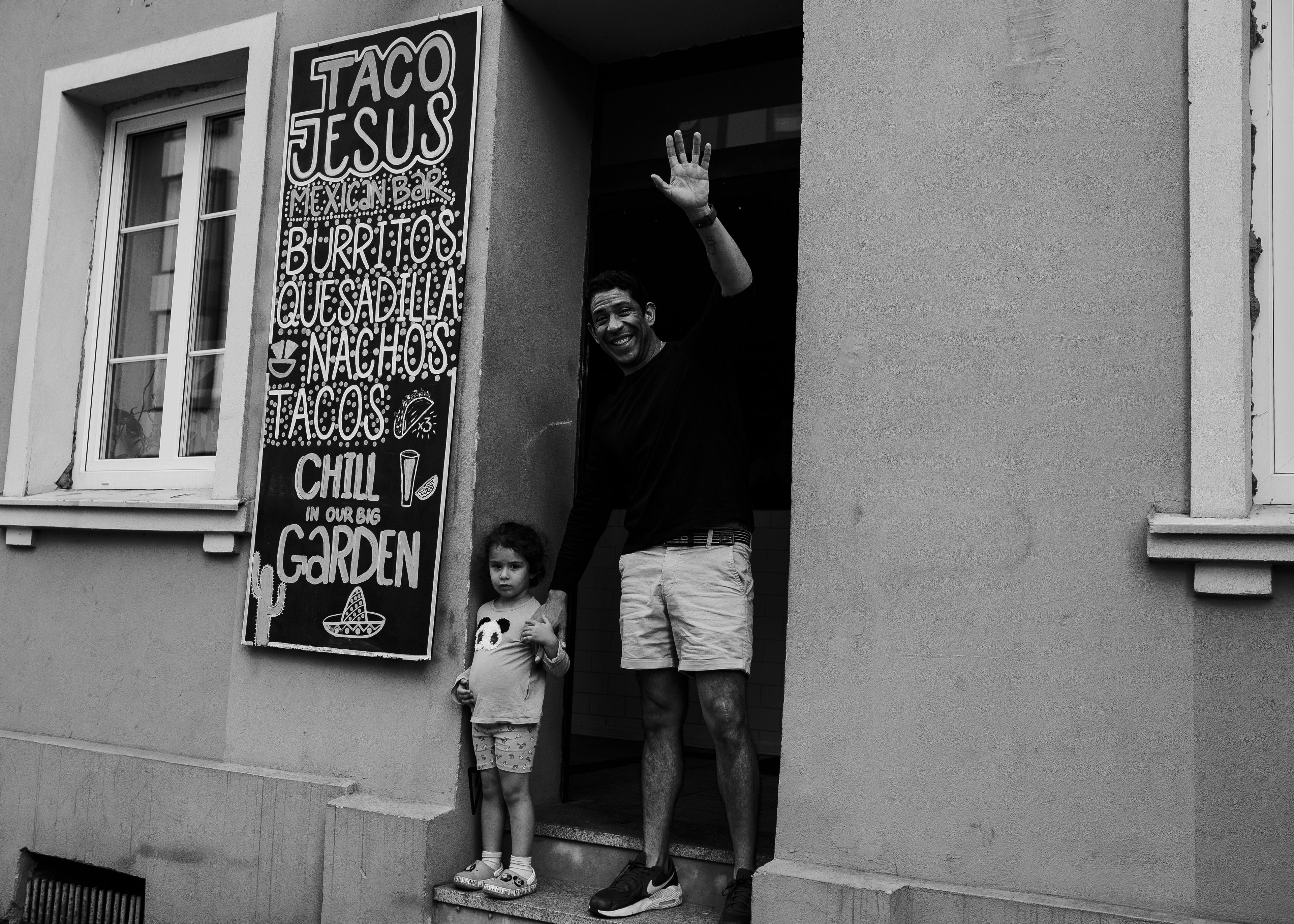 młody czarnoskóry mężczyzna stoi w drzwiach kamienicy, trzyma za rękę małą dziewczynka uśmiecha się i pozdrawia ręką