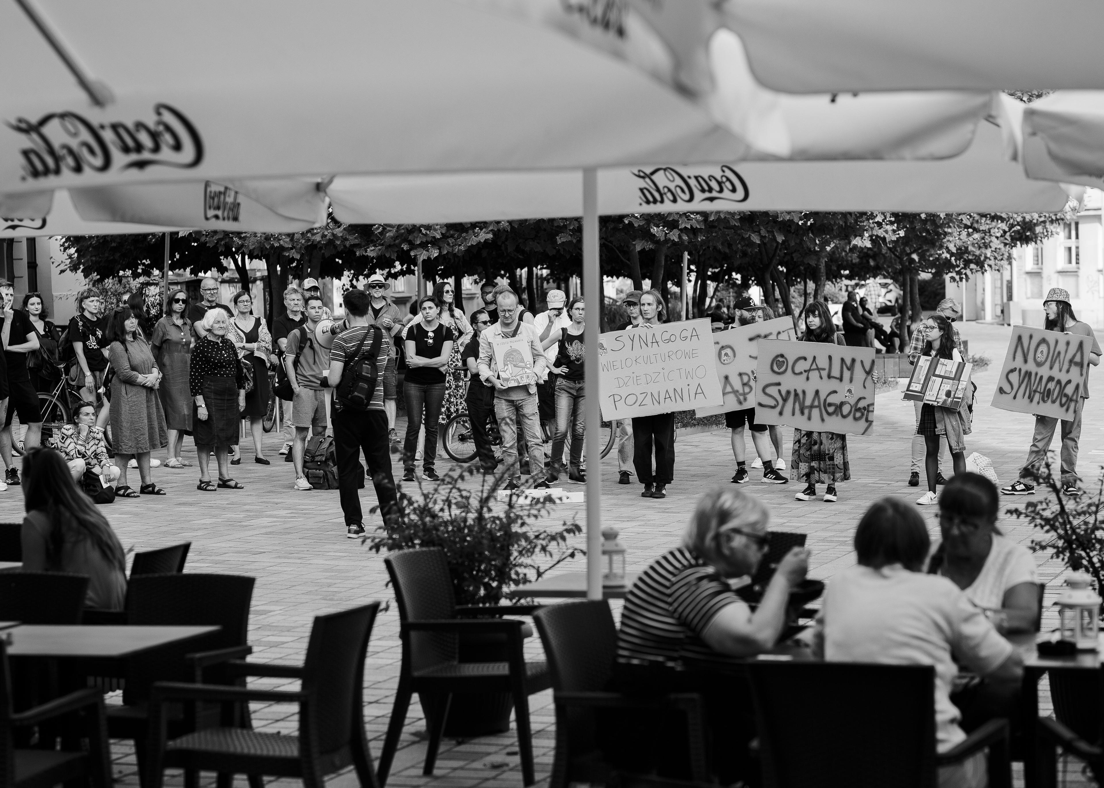 widok na manifestację uliczną spod parasoli ogródka miejskiej kawiarni. na parasolach napis cocacola, w głębi ludzie z hasłami w obronie poznańskiej synagogi
