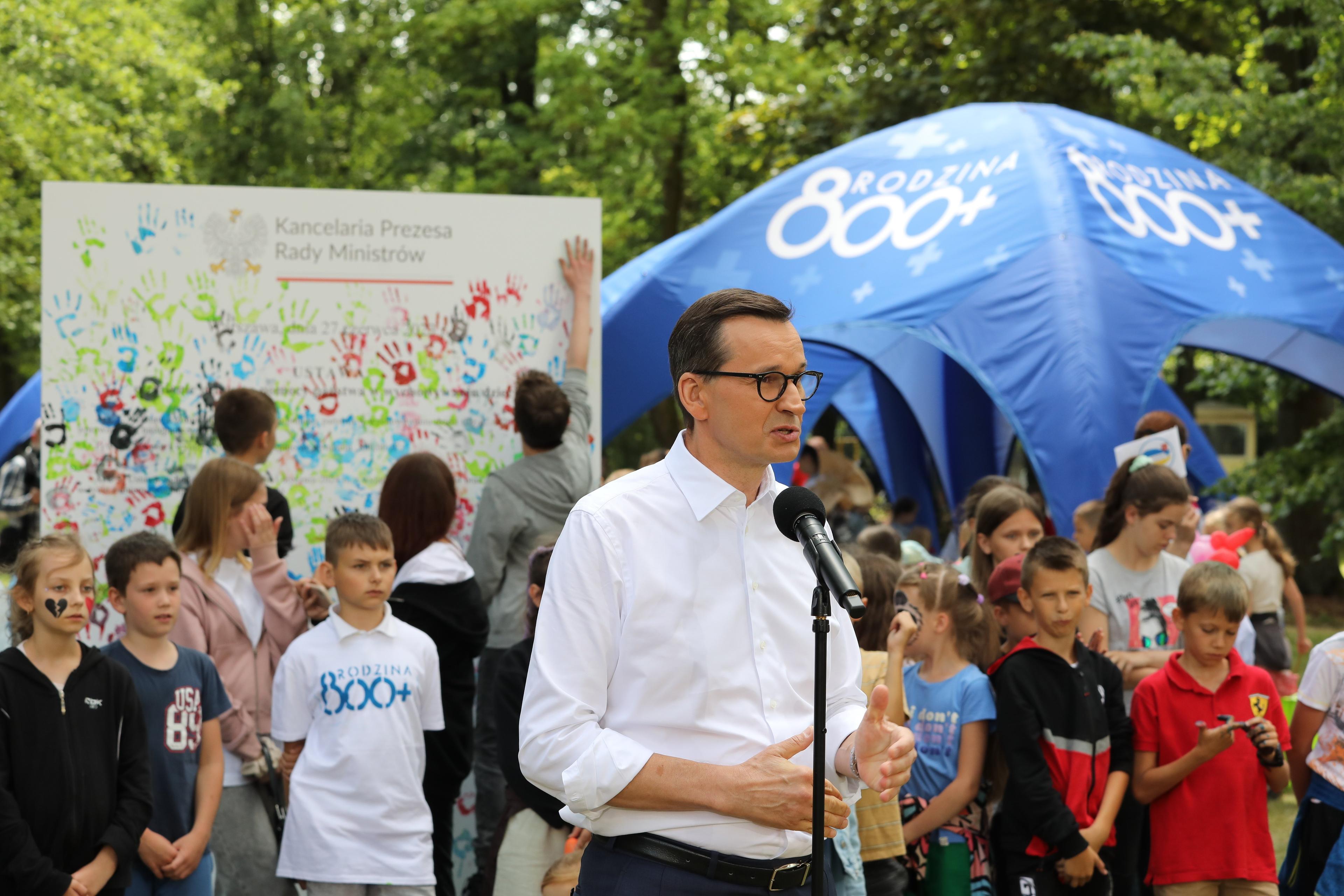 Premier Mateusz Morawiecki przemawia do mikrofonu na tle niebieskiego namiotu z napisem "rodzina 800+"