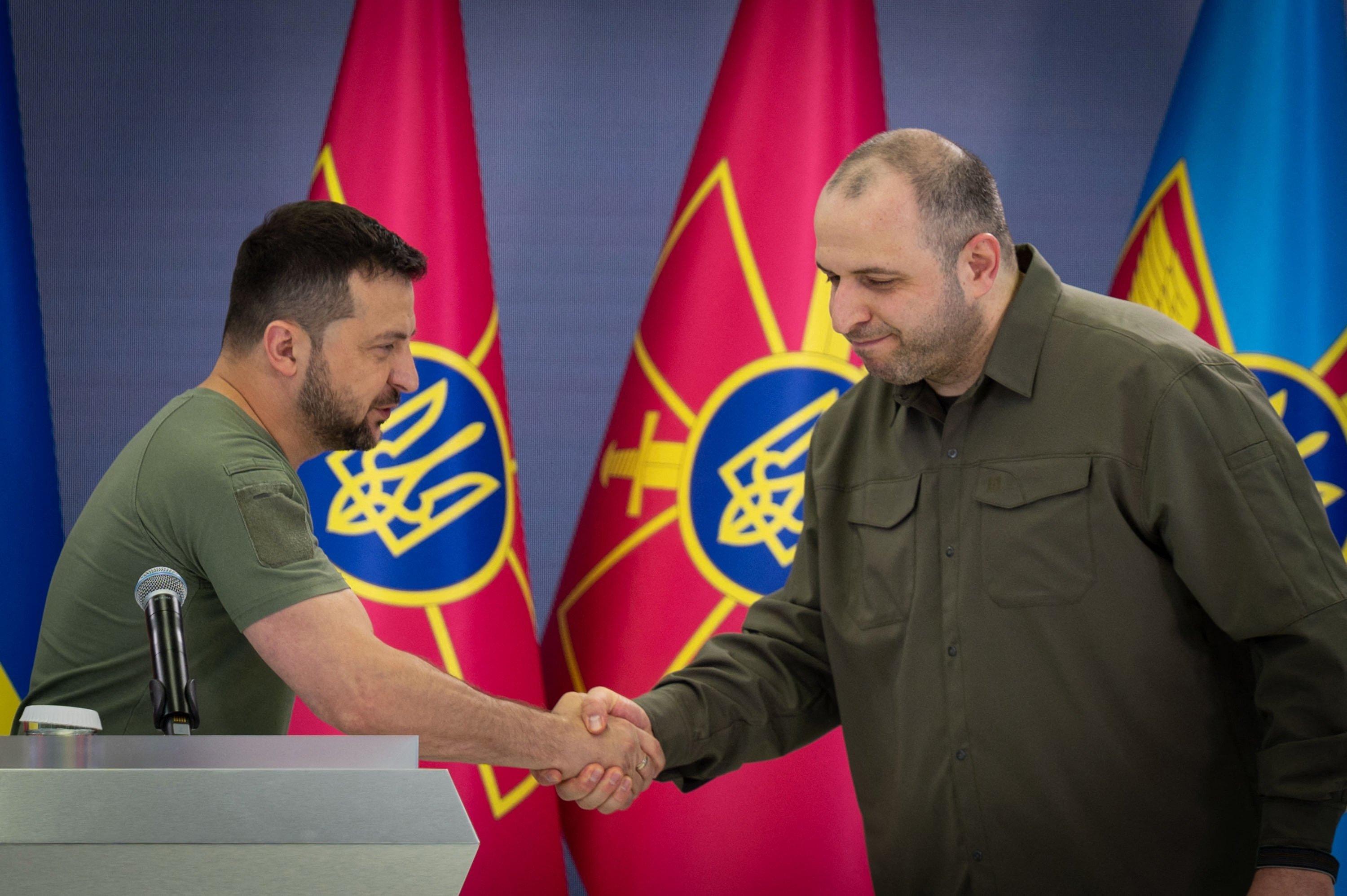 Prezydent Wołodymyr Zełenski ściska rękę ministra obrony Rustema Umarowa na podium z flagami