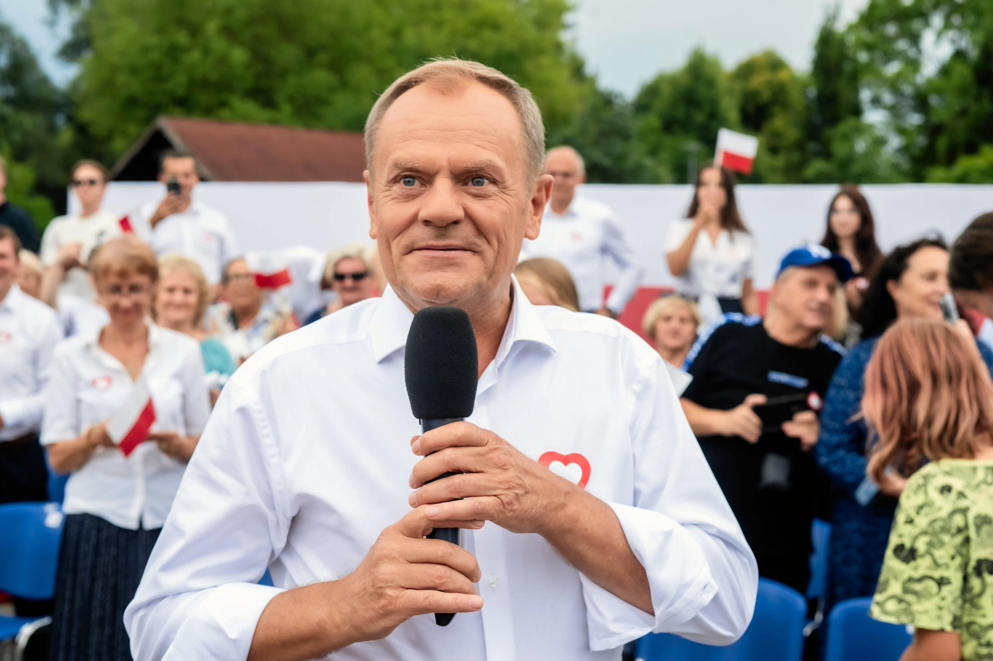 Uśmiechnięty Donald Tusk w białej koszuli stoi na tle wyborców, trzymając mikrofon.