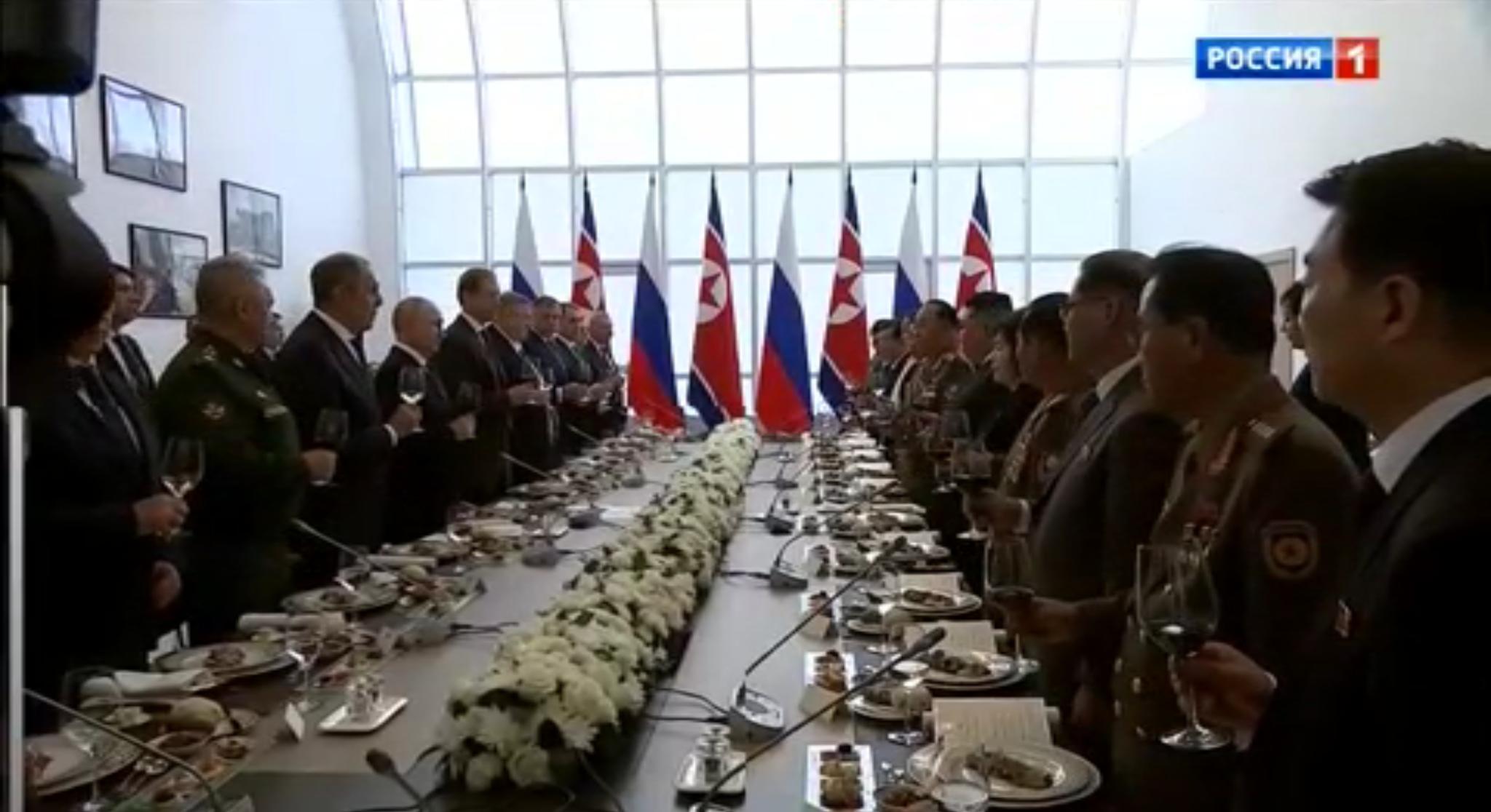 Męzczyźni stoją z kieliszkami przy suto zastawionym stole. W tle flagi Rosji i Korei Północnejwznoczą toast