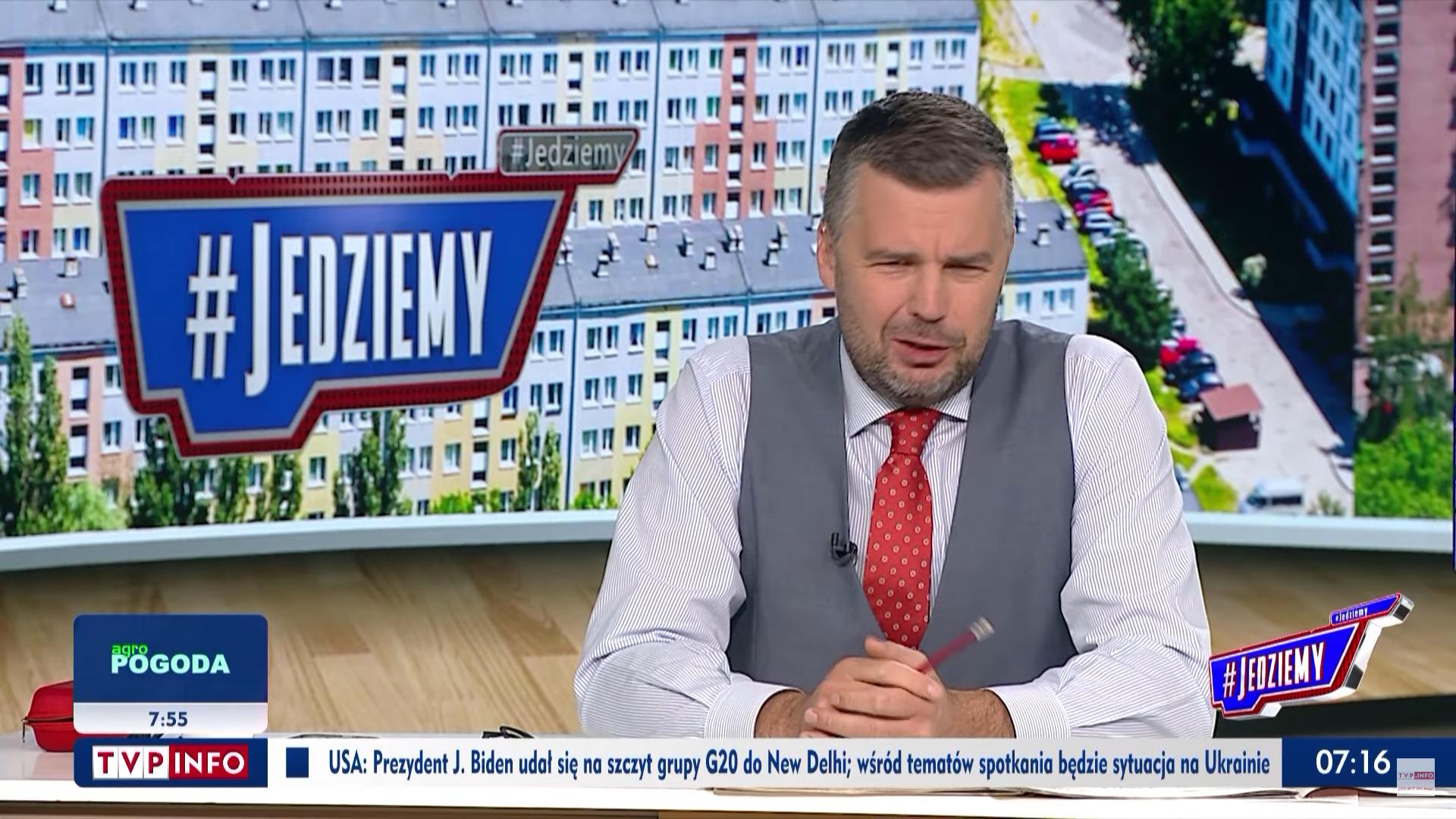 Michał Rachoń z TVP w szarej kamizelce i czerwonym krawacie przemawia do kamery na tle napisu #Jedziemy