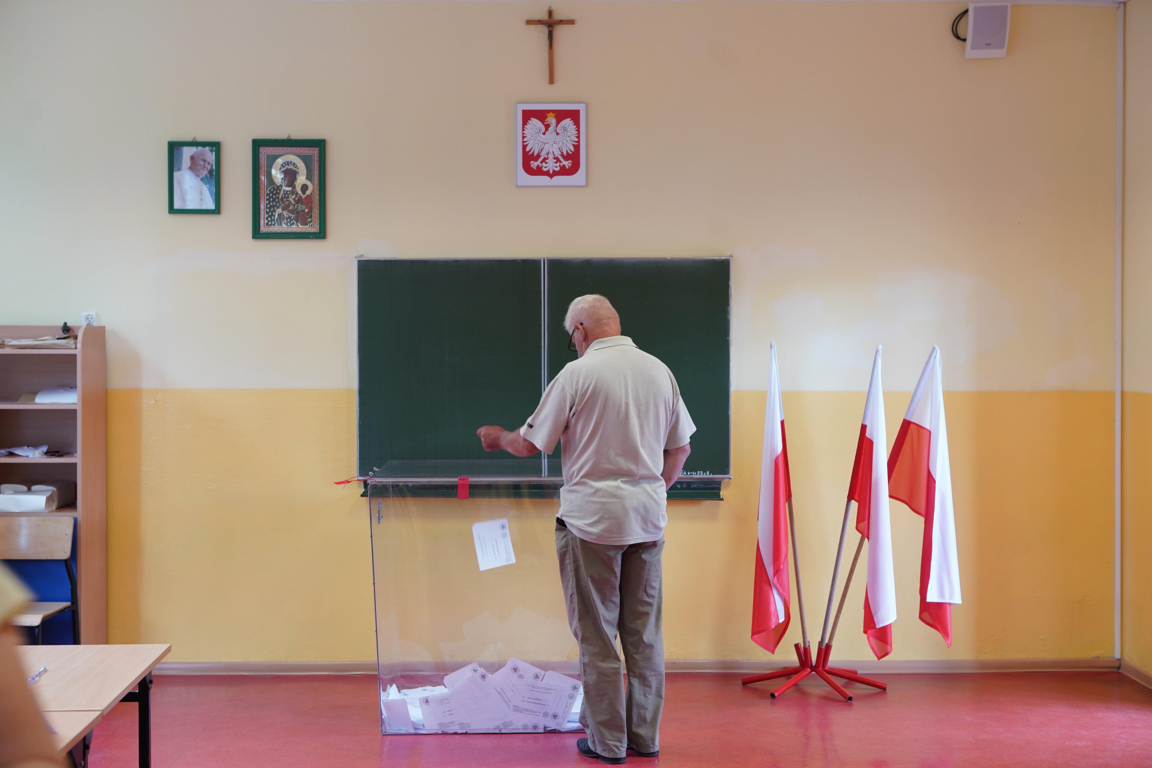 Na zdjęciu widać szkolną klasę z godłem, tablicą i krzyżem na ścianie. Mężczyzna w starszym wieku wrzuca kartę do głosowania do przezroczystej urny wyborczej. Obok stoją trzy polskie flagi