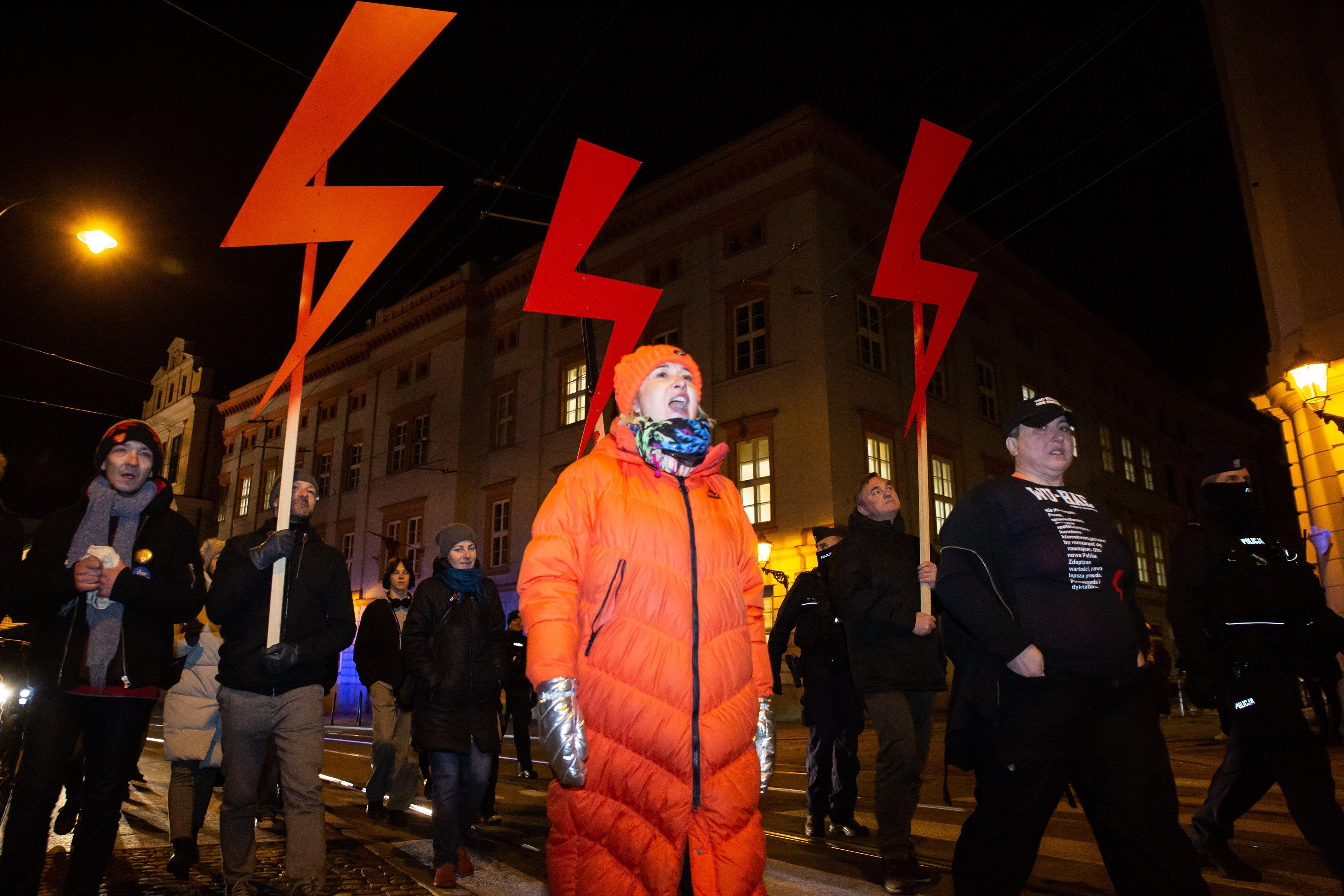 Kobieta w pomarańczowej kurtce stoi na ulicy, nad nią błyskawice jako symbol Strajku Kobiet
